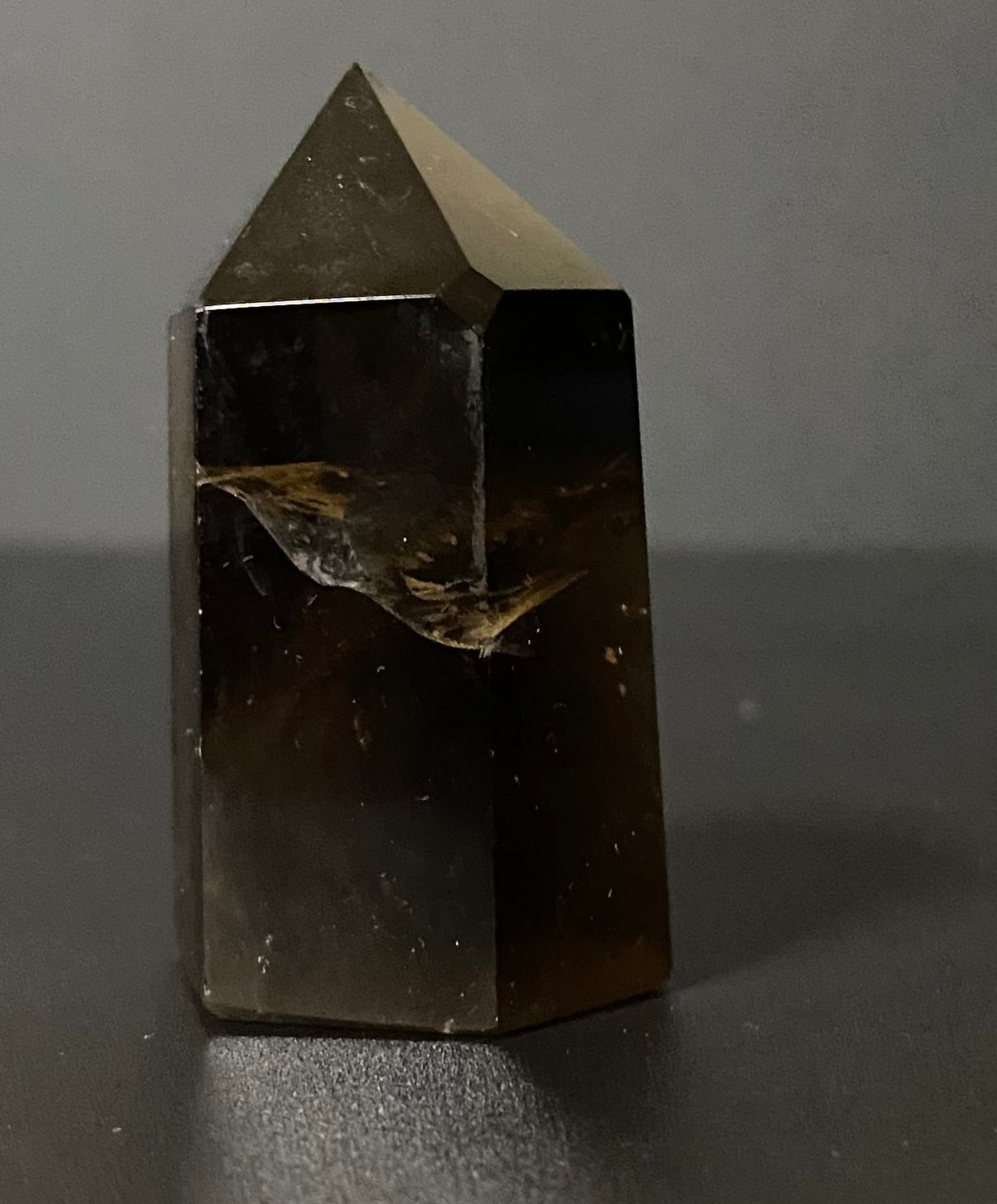 A natural smoky quartz crystal point with a hexagonal shape, showing translucent brown and amber tones with internal inclusions, standing on a dark surface against a neutral gray background.