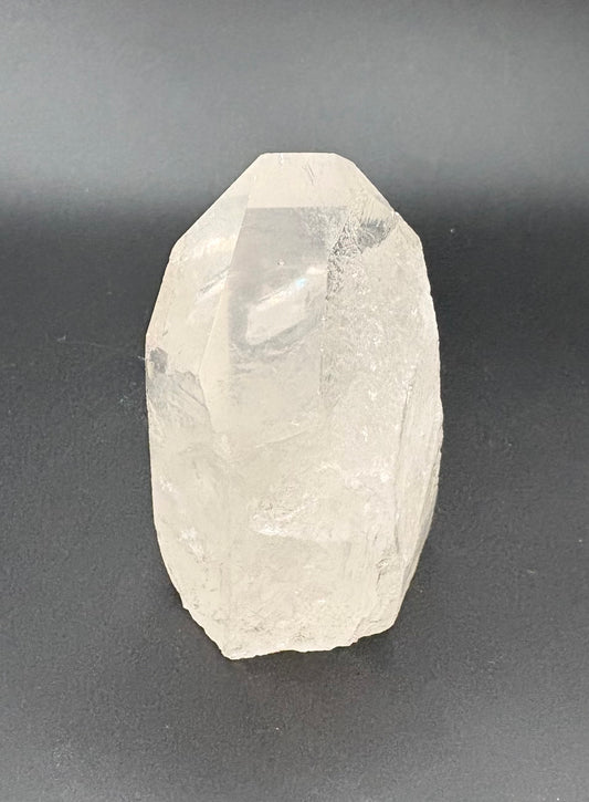 A raw, faceted clear quartz crystal point standing upright on a dark gray surface.