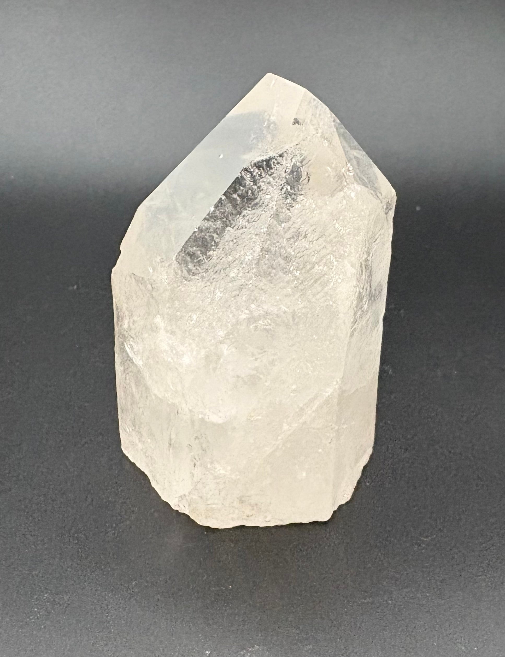 A raw, unpolished clear quartz crystal point with natural rough edges and internal inclusions, photographed on a dark gray background.