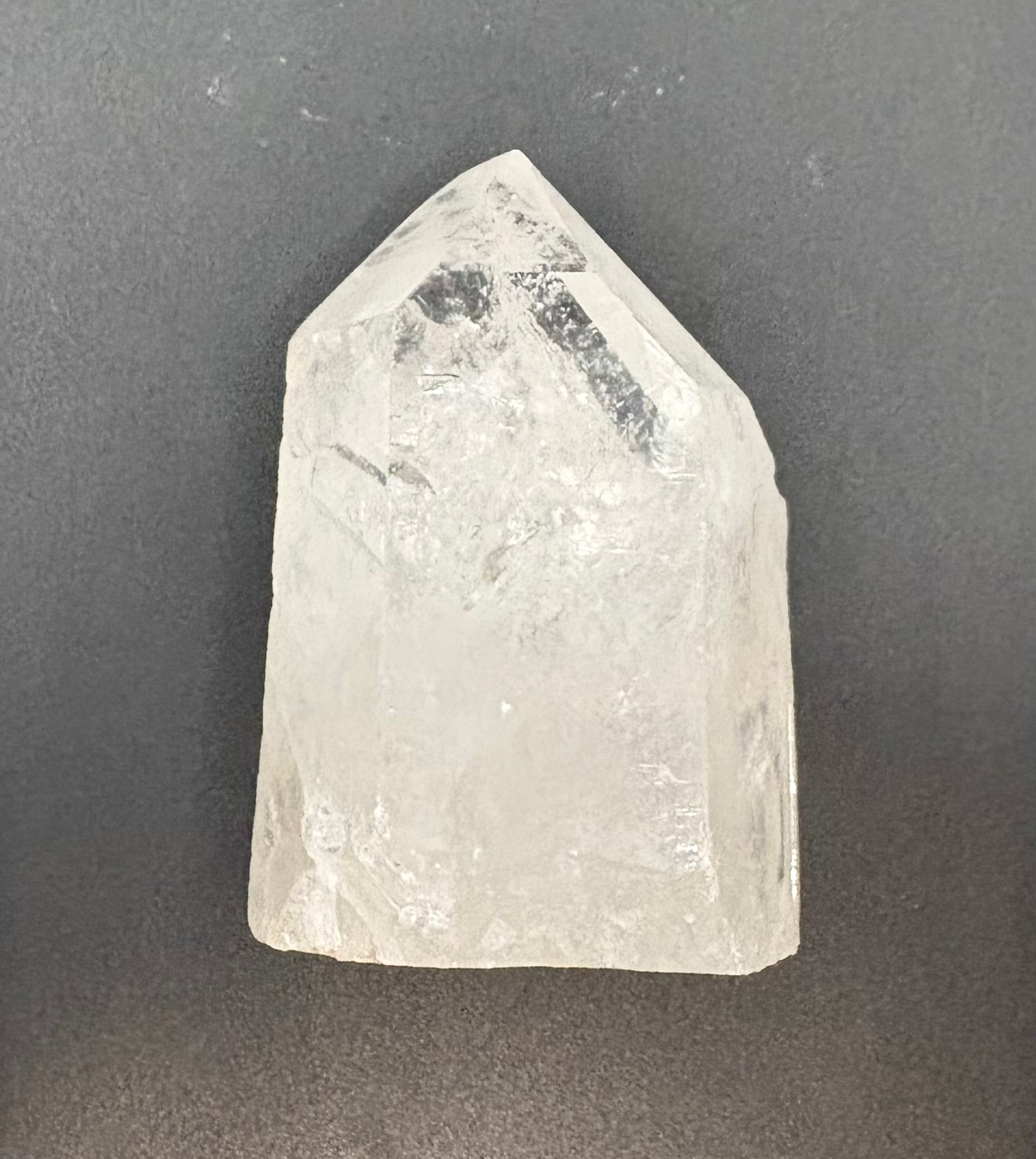 A raw, unpolished clear quartz crystal point with natural inclusions, displayed on a dark gray textured background.