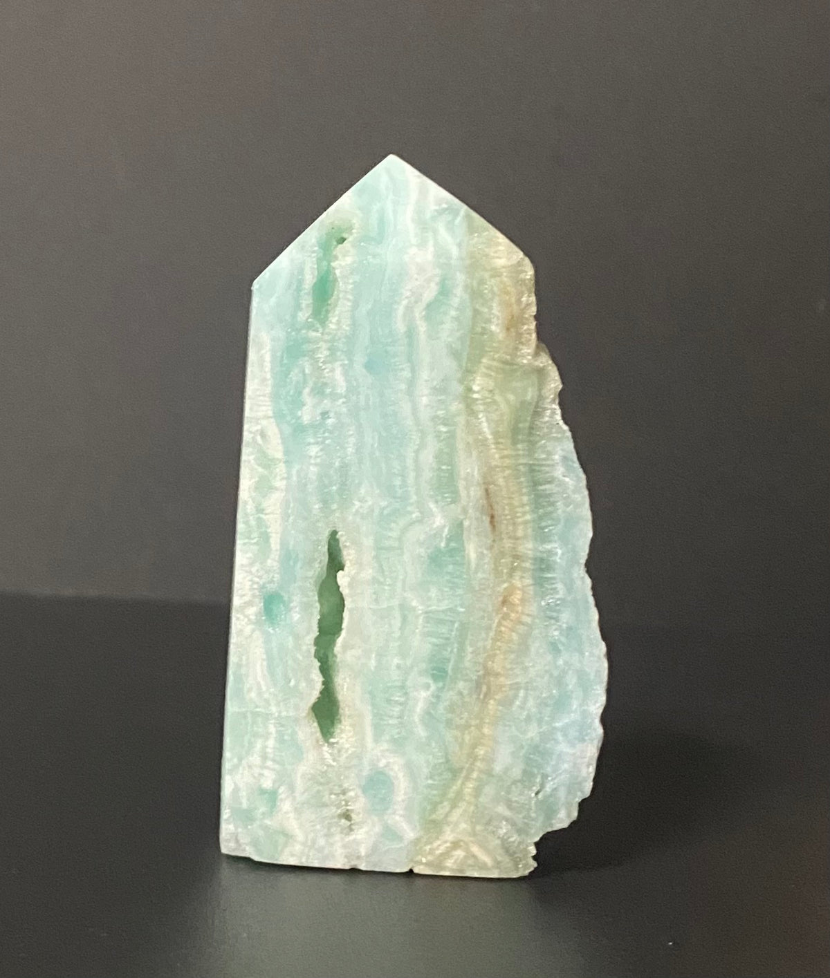 A raw Calcite crystal point with natural green and white banded patterns, standing upright on a dark surface against a dark gray background.