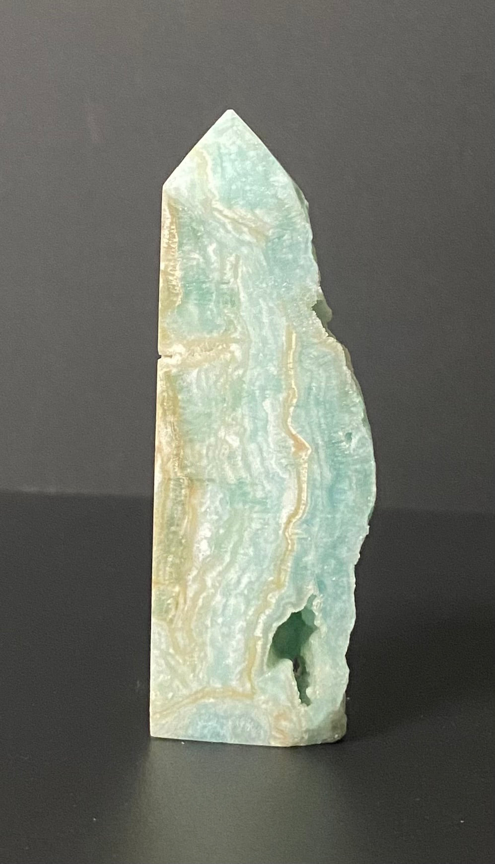 A polished natural mineral point or crystal tower, featuring light aqua green and cream marbling, standing upright on a dark gray surface.