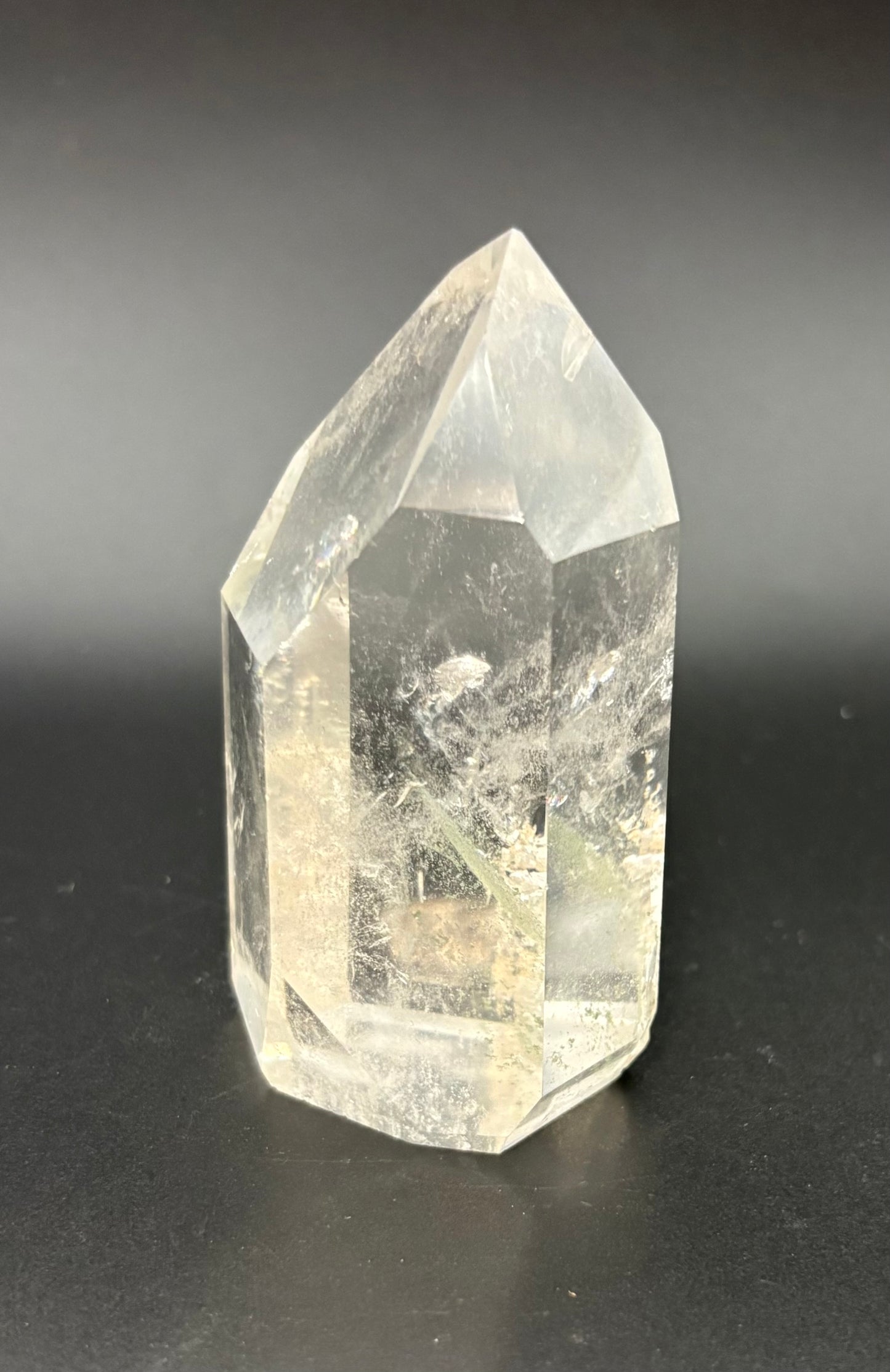 A natural clear quartz crystal point with internal inclusions, standing on a dark reflective surface against a dark gray background.