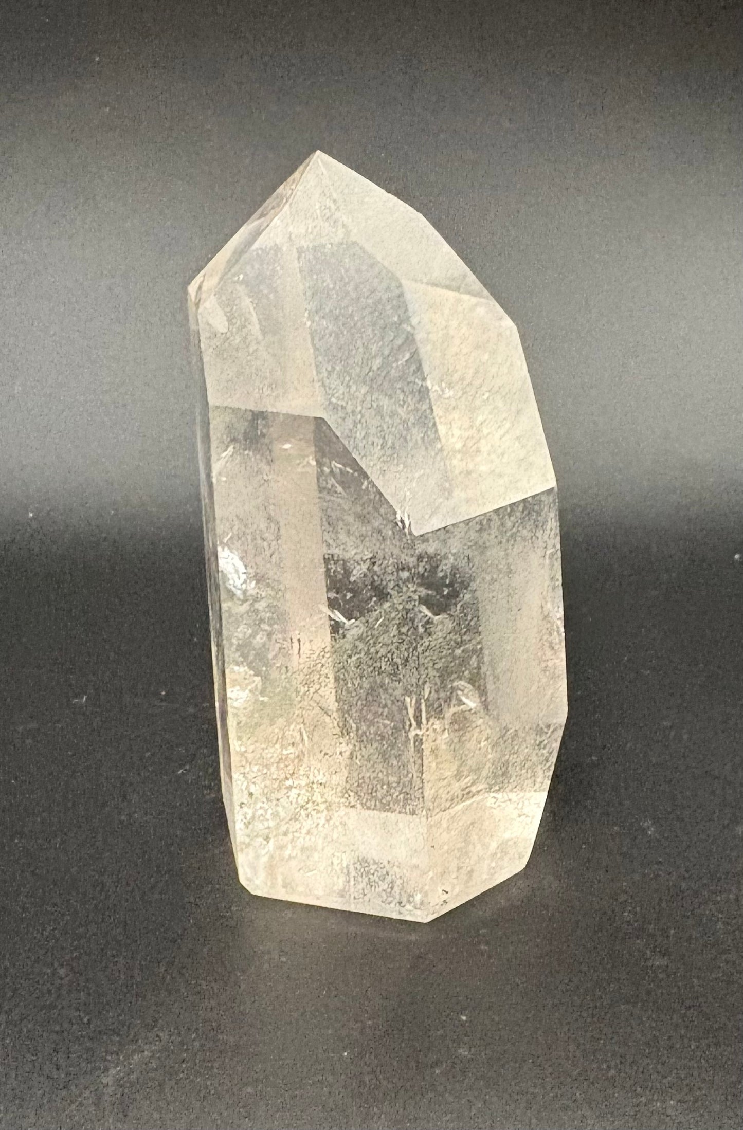 A natural clear quartz crystal point tower with internal inclusions, standing on a dark gray surface against a dark gray background.