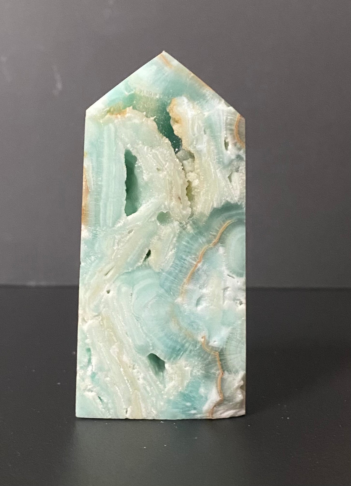 A pointed rectangular polished stone or crystal with marbled aqua, white, and pale green veining, standing upright on a dark surface against a dark gray background.