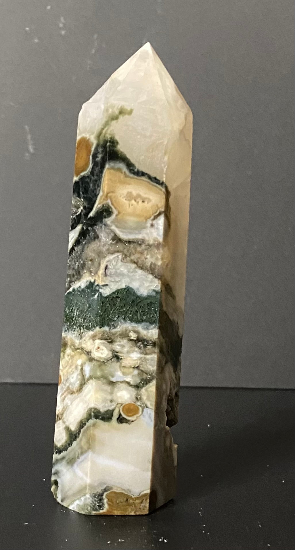 A natural pointed crystal Tree Agate tower or mineral spire with marbled patterns of white, gray, beige, and green, standing on a dark surface against a dark gray background.