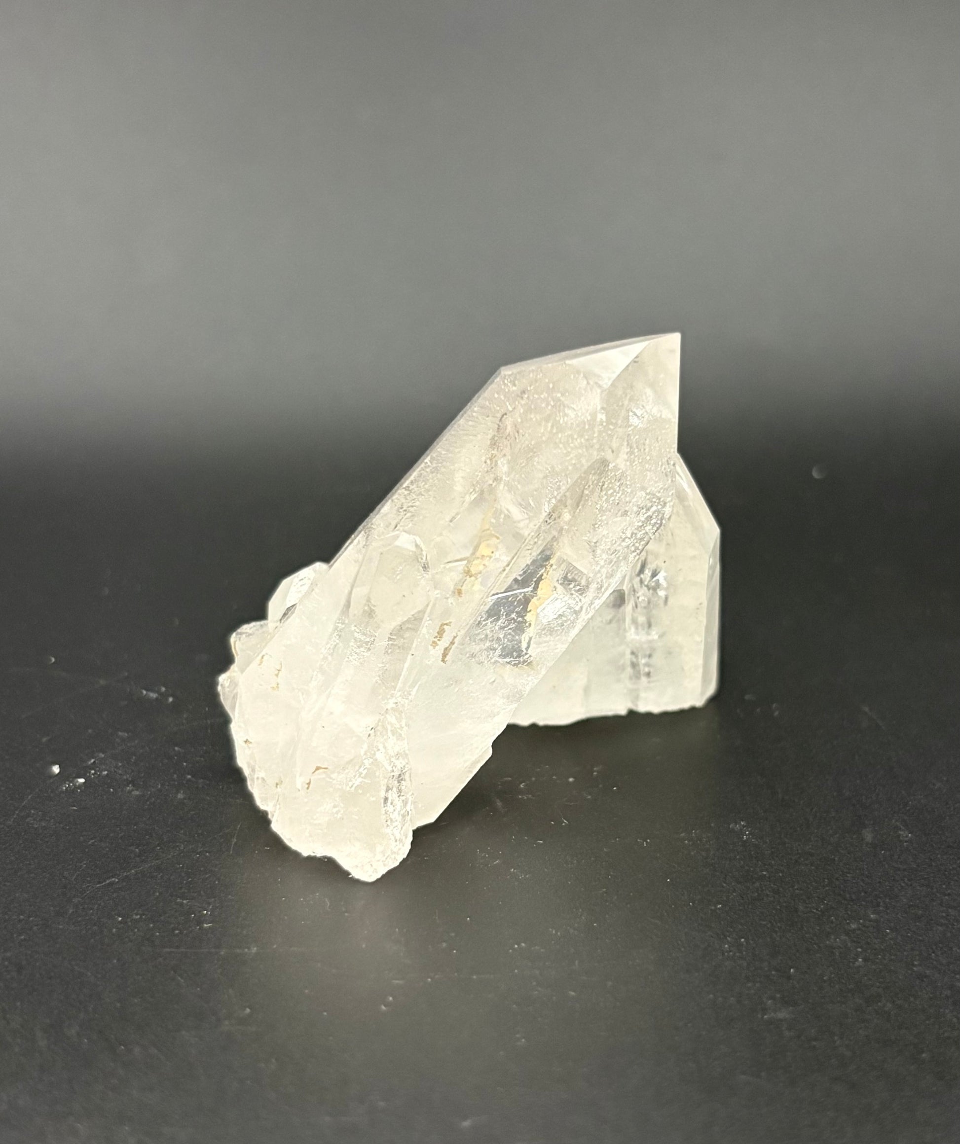 A raw, unpolished clear quartz crystal cluster with natural geometric facets, resting on a dark gray surface against a dark gray background.