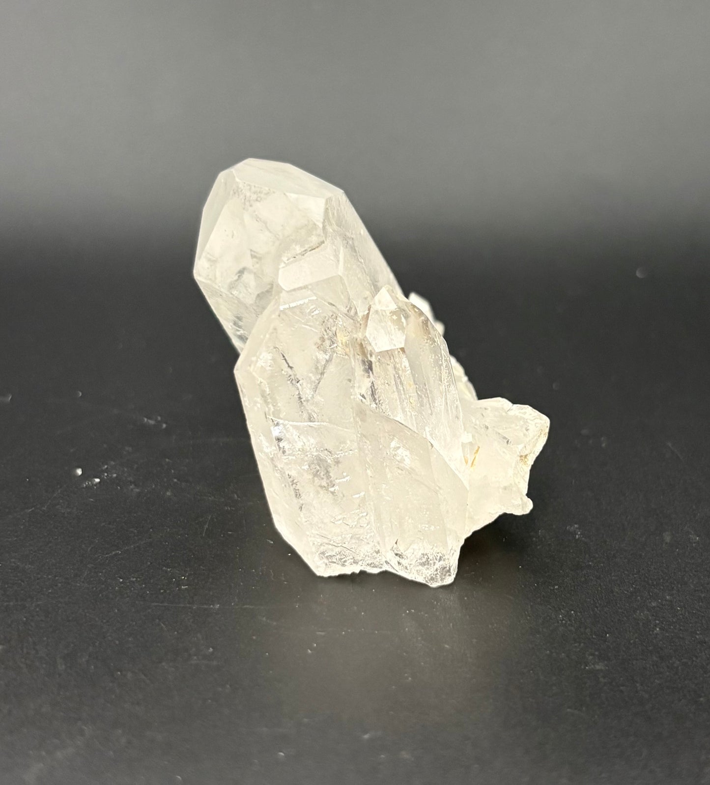 A raw, unpolished clear quartz crystal cluster with sharp, faceted edges, resting on a dark gray surface against a dark gray background.