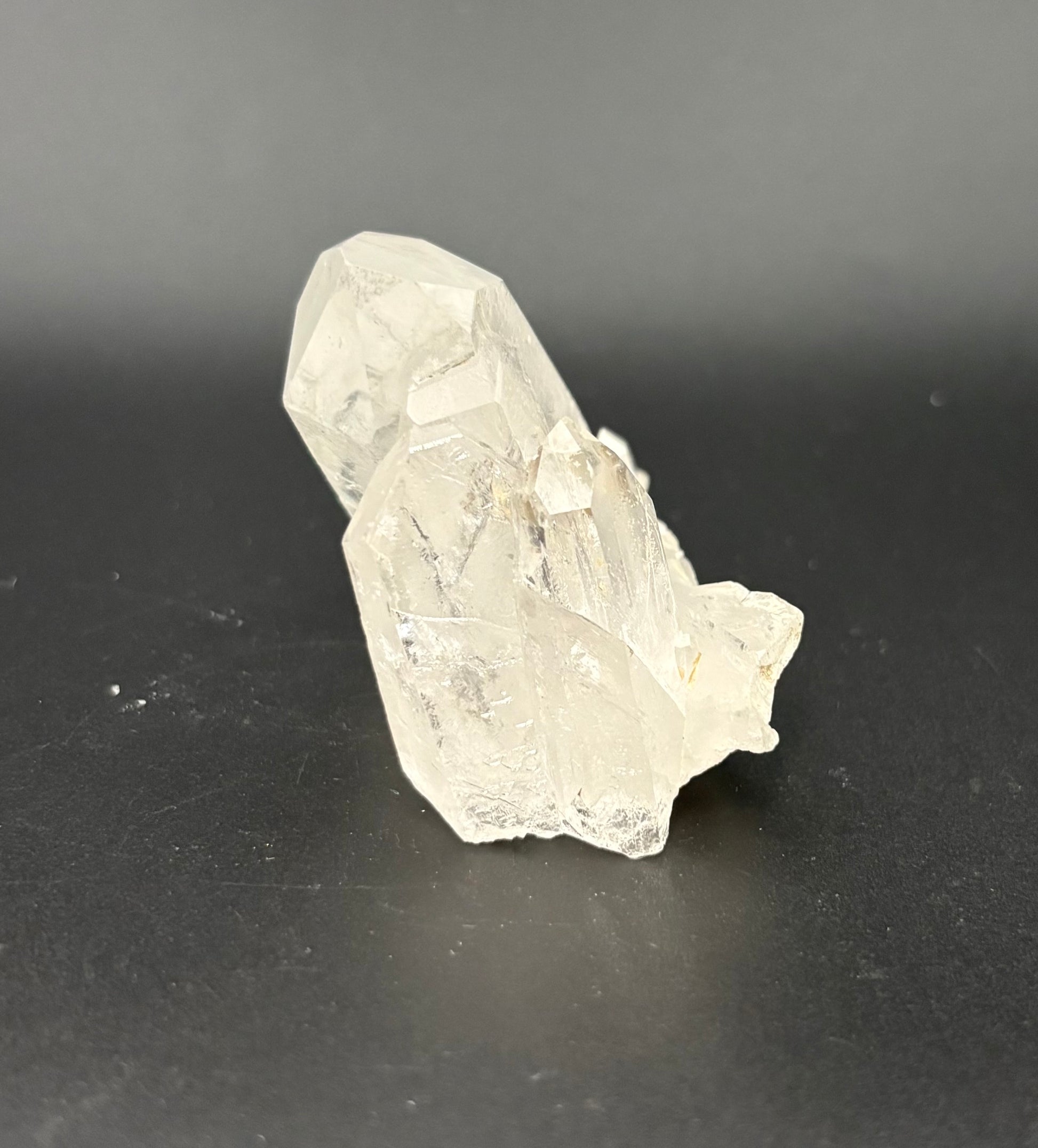 A raw, unpolished clear quartz crystal cluster with sharp, faceted edges, resting on a dark gray surface against a dark gray background.
