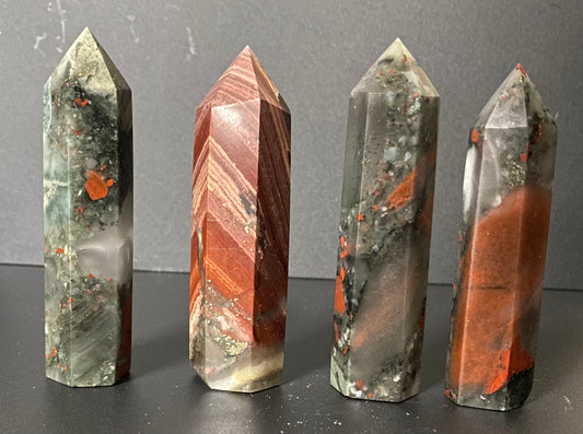 Four polished crystal points Bloodstone with natural banding and inclusions, displayed upright on a dark surface against a gray background.