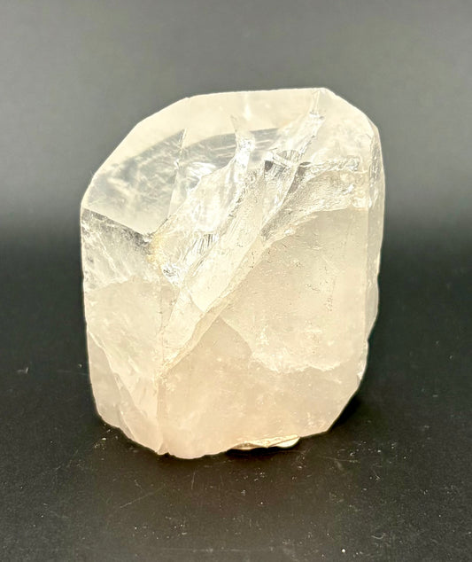 A rough, unpolished natural quartz crystal with a pale, milky appearance, standing upright on a dark gray surface.