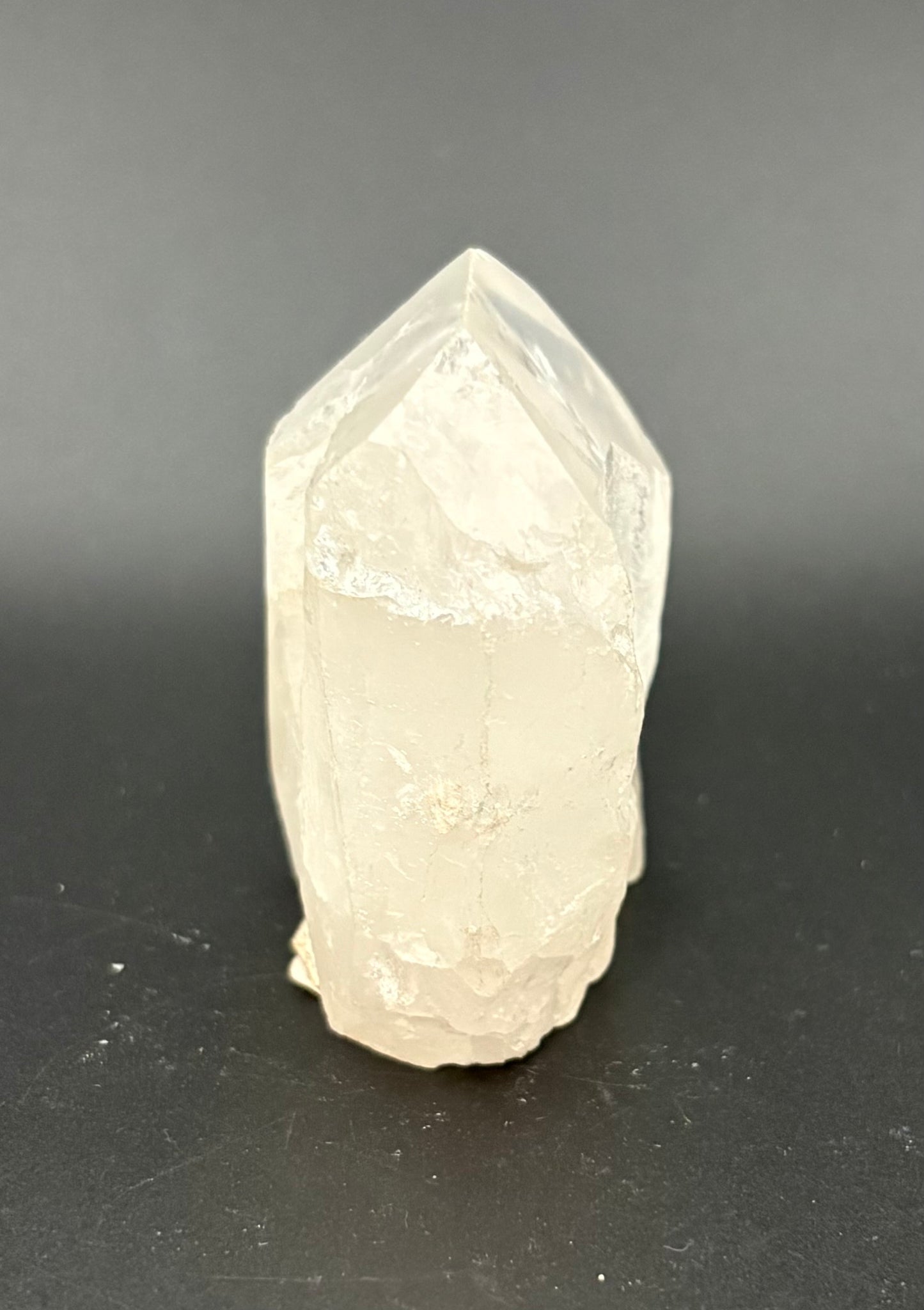 A natural white quartz crystal point with rough, unpolished facets, standing upright on a dark gray surface.