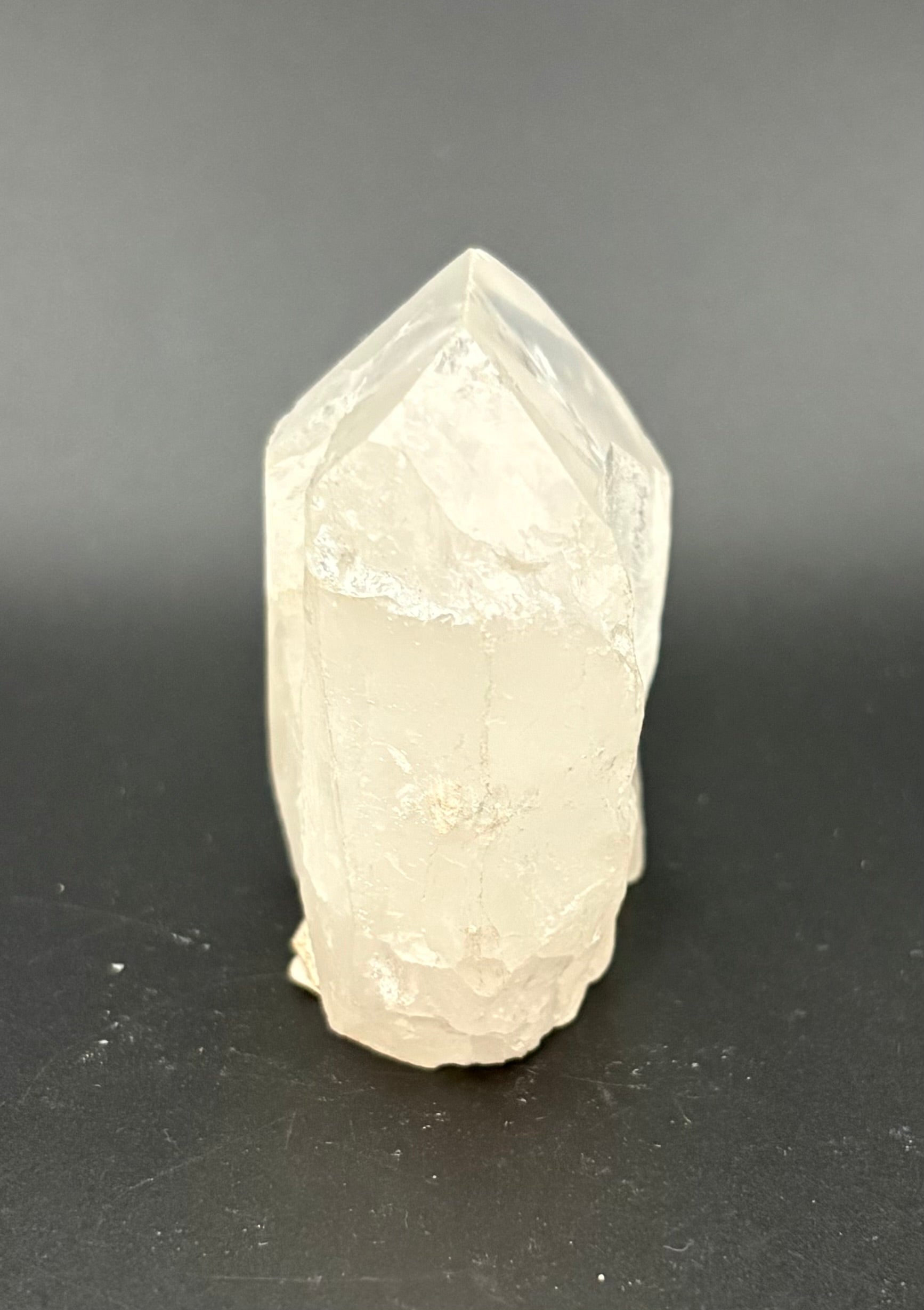 A natural white quartz crystal point with rough, unpolished facets, standing upright on a dark gray surface.