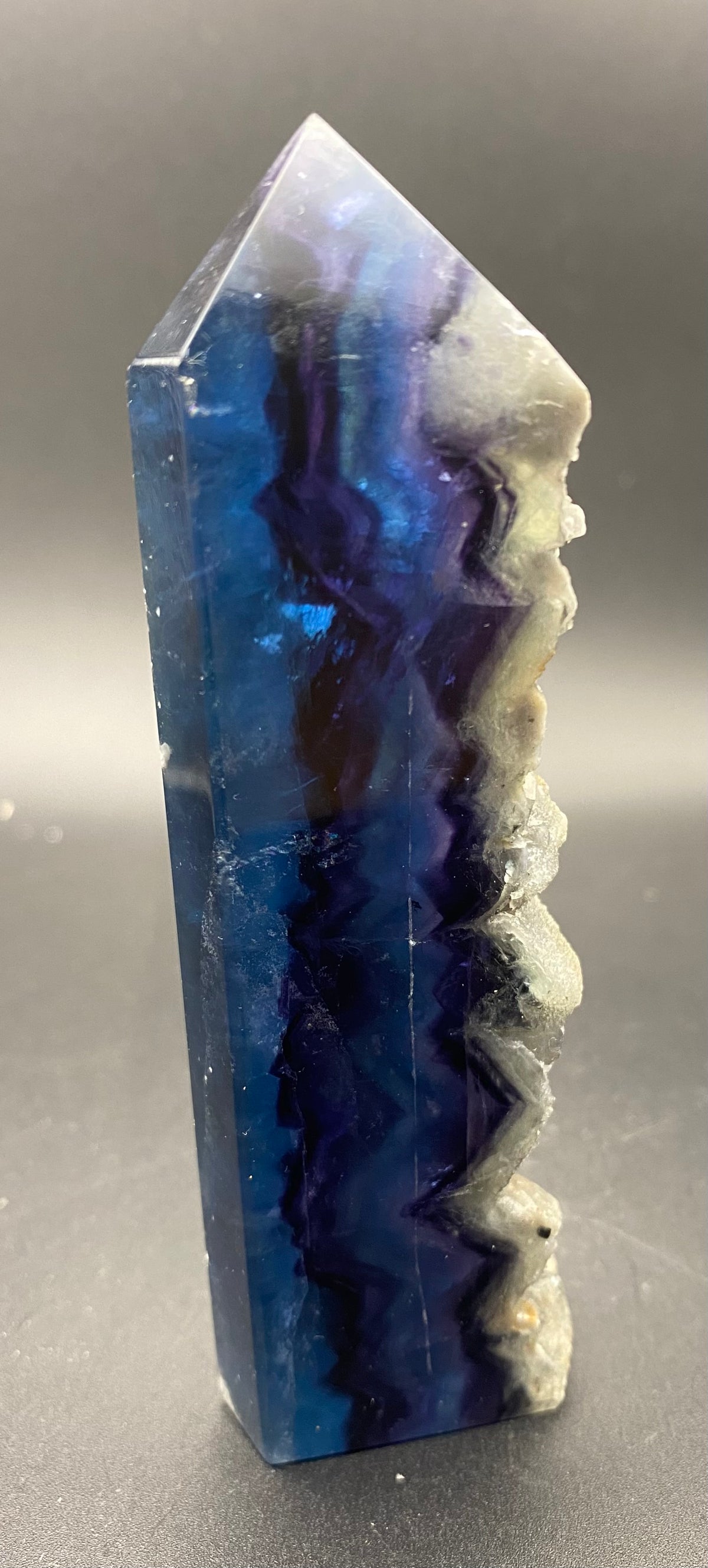 A polished fluorite natural crystal tower with a pointed top, featuring a marbled pattern of deep blue, indigo, and white with a rough, raw edge on the right side, set against a dark gray background.