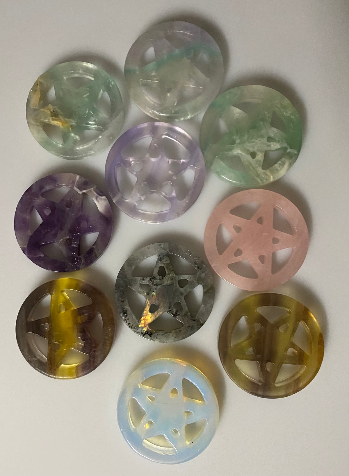 Nine circular translucent gemstone beads with carved pentacle/star cutouts, arranged on a plain light background. The beads show different colors including green, purple, pink, gray, yellow, and light blue.