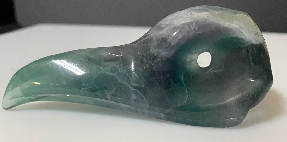 A carved fluorite green raven skull figurine made of polished green and gray stone, featuring a hollow eye socket, resting on a white surface against a dark gray background.