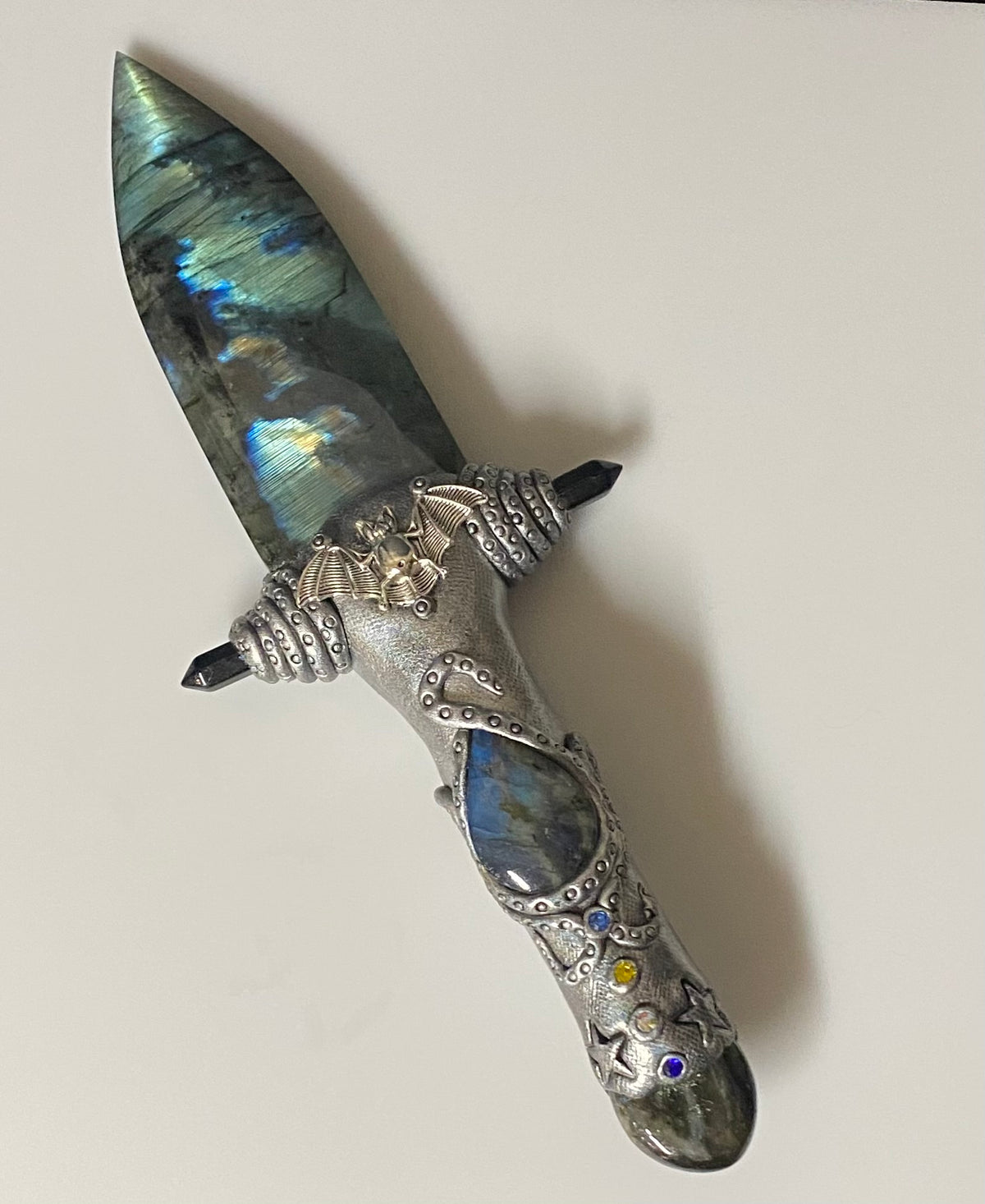 A decorative dagger with a polished iridescent labradorite blade and an ornate silver-toned hilt featuring gemstone accents, photographed on a plain light background.