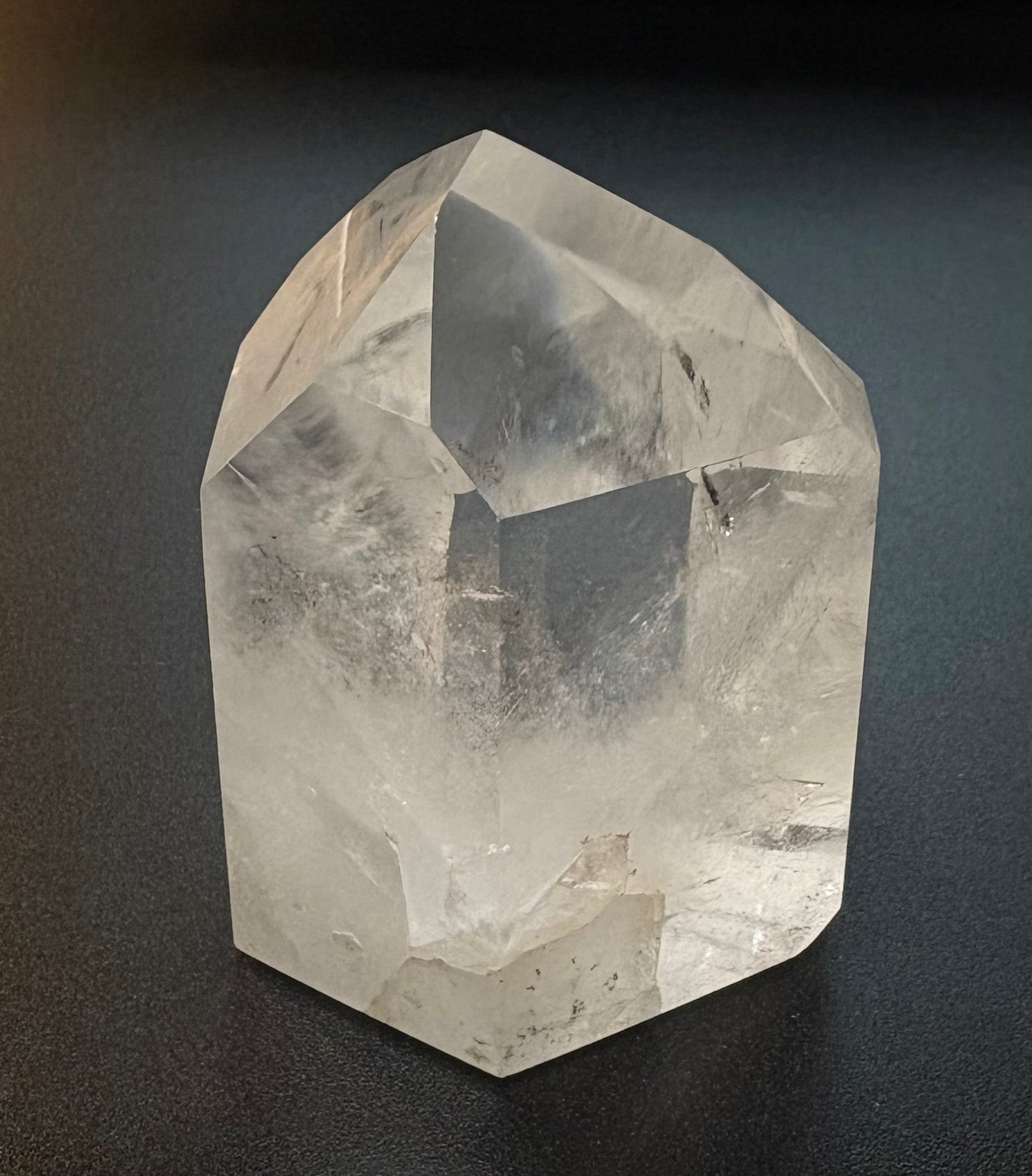A natural clear quartz crystal cluster with rough, faceted edges, displayed on a dark textured surface.