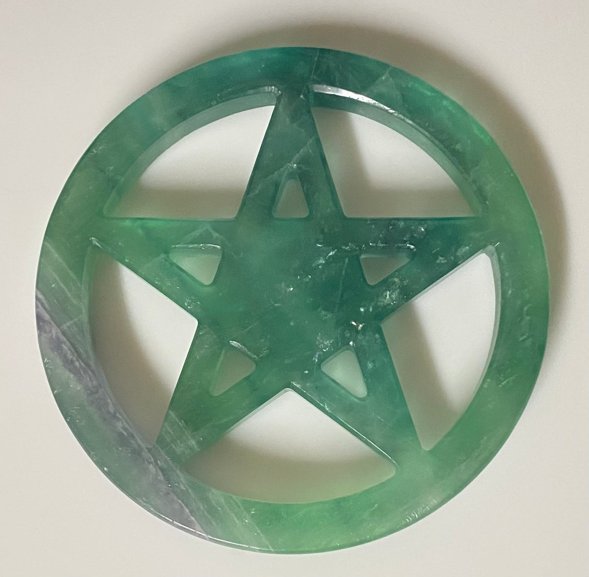 A circular fluorite pentacle featuring a translucent green color with natural stone texture and a polished finish, shown on a light beige background.