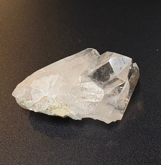 A raw, rough crystal cluster with clear faceted edges and natural imperfections, resting on a dark, textured surface.