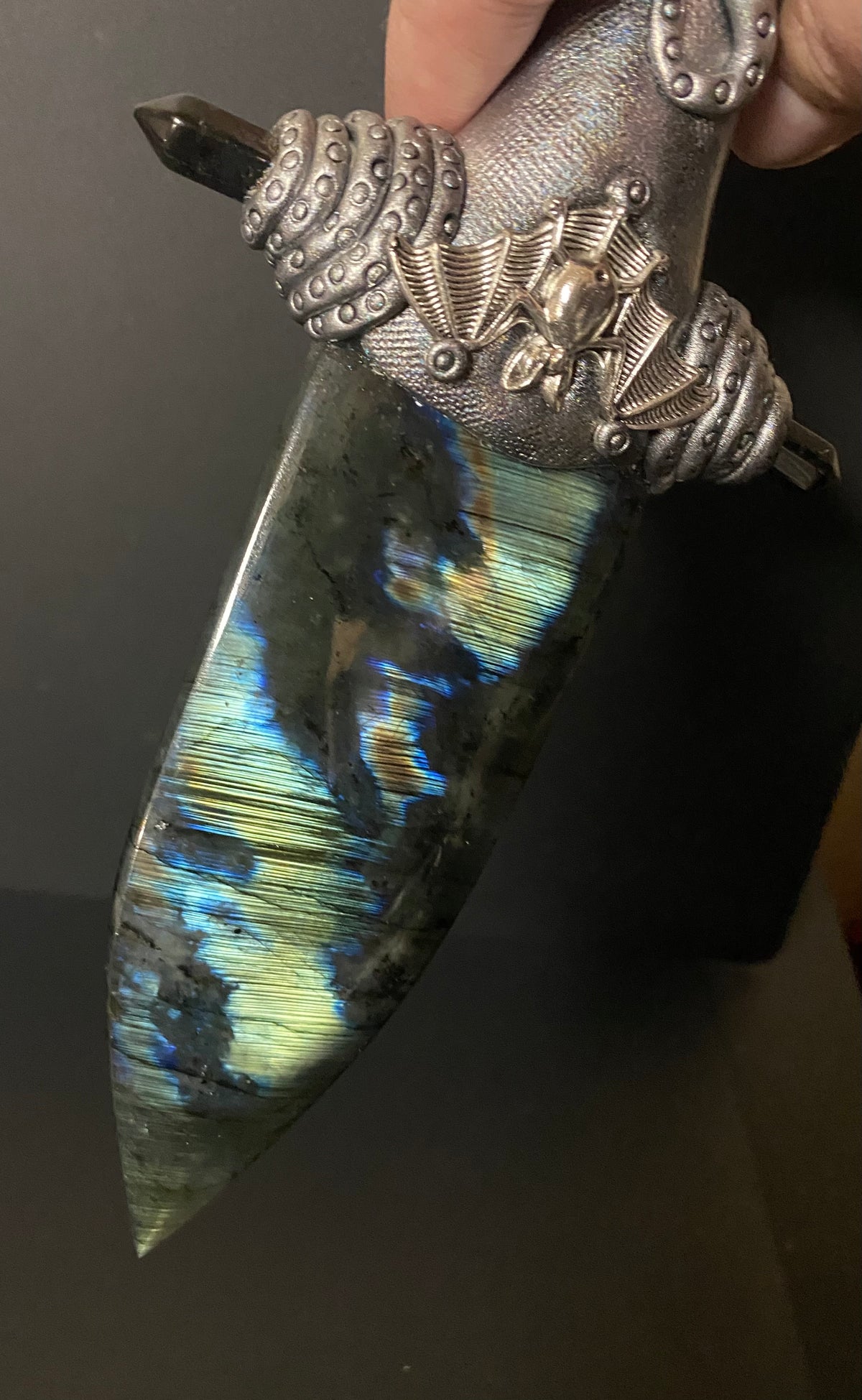 A hand holds a decorative dagger with a polished labradorite iridescent blade showing blue, green, and gold flashes, a textured silver hilt, and a dark pommel against a dark background.