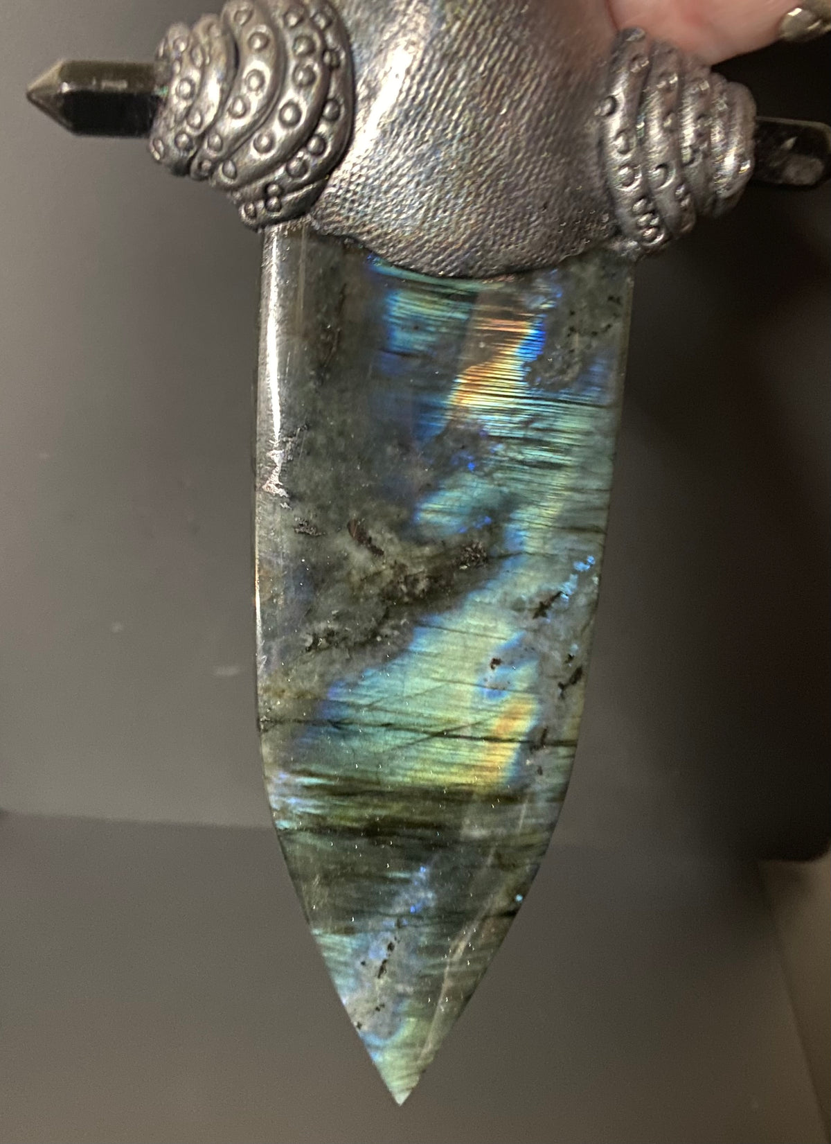 A close-up of a decorative dagger or knife with a polished iridescent labradorite blade and an ornate silver-toned metal hilt, held against a dark gray background.
