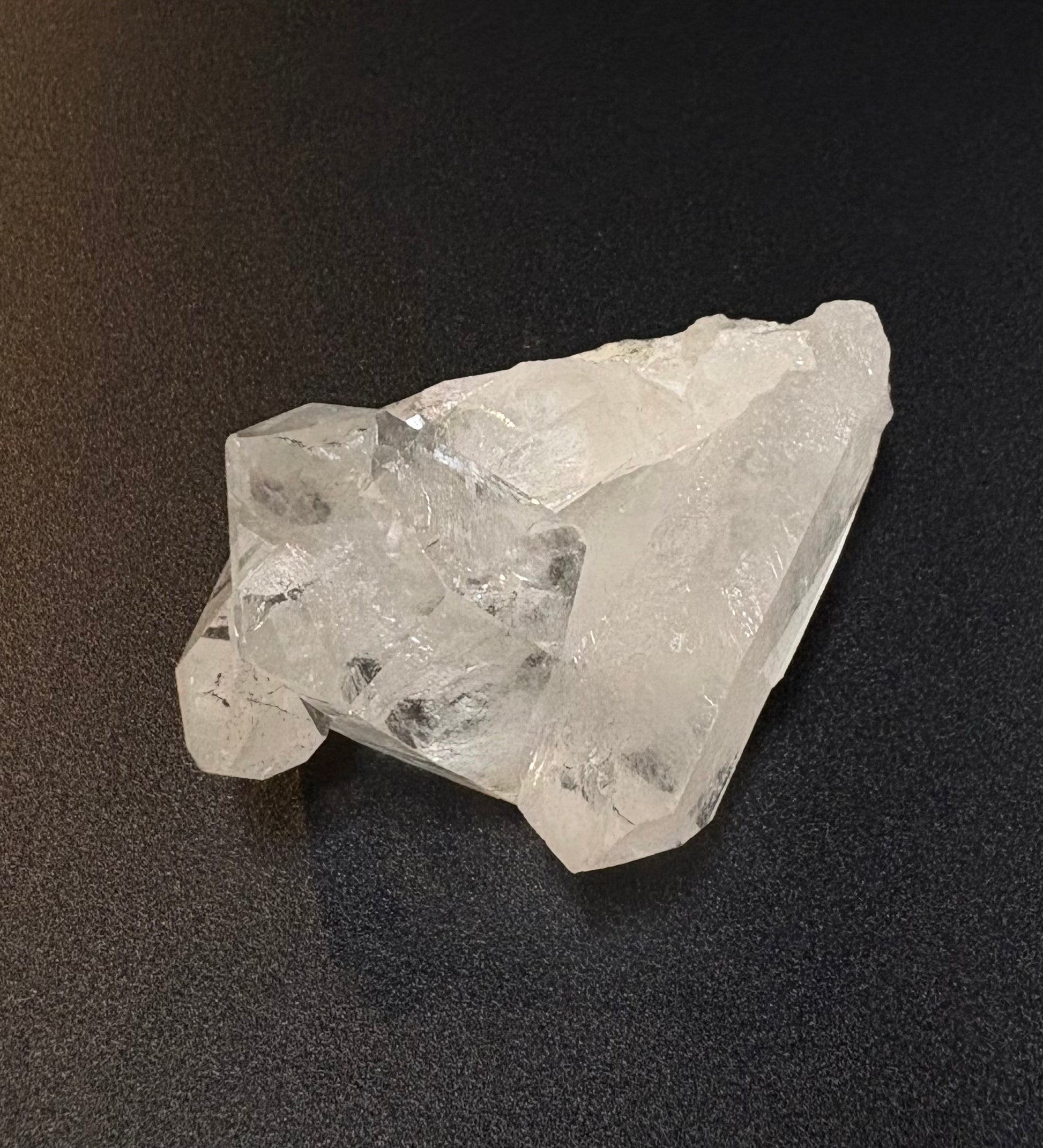 A raw, unpolished clear quartz crystal cluster with visible natural facets and impurities, resting on a dark textured surface.