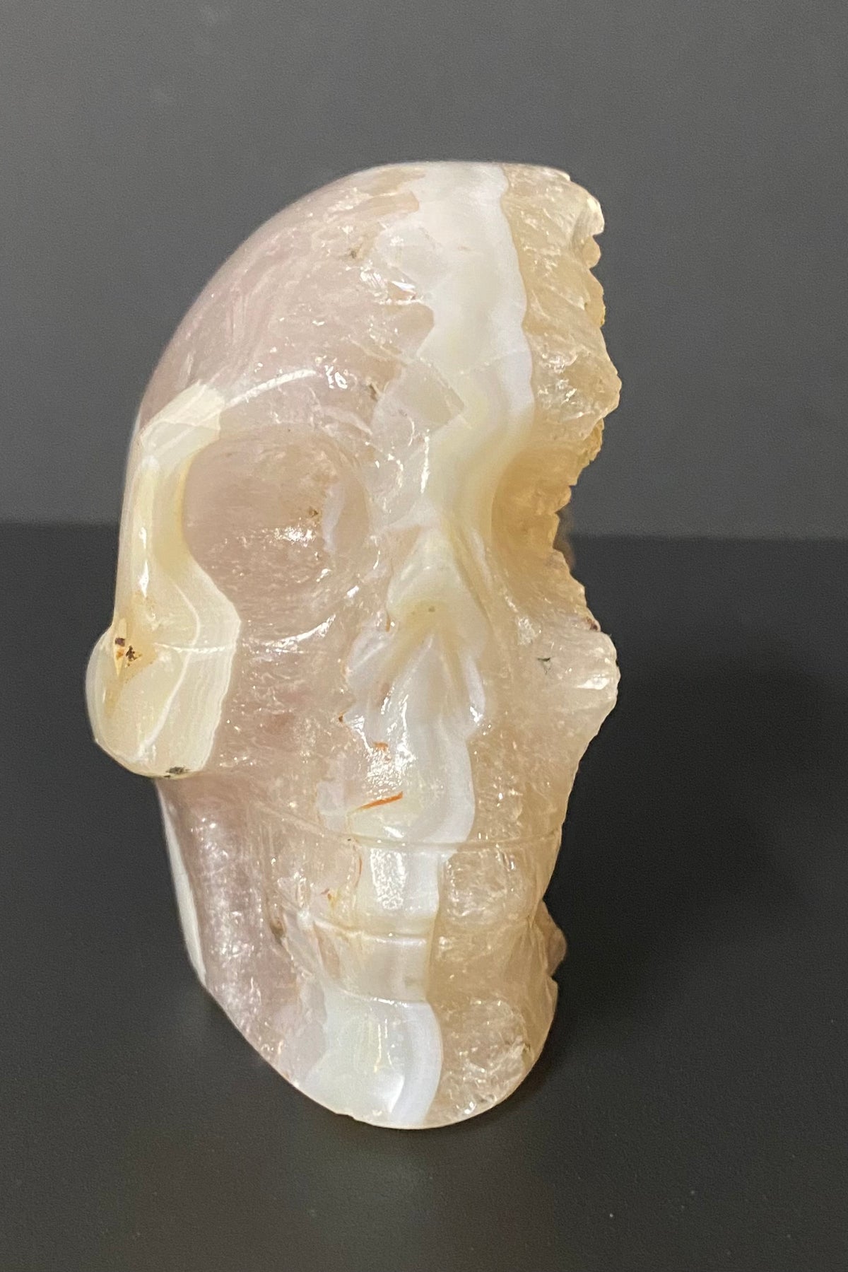 A carved stone skull sculpture made from flower agate and quartz, showing natural banding in milky white and pale peach-beige tones, photographed against a dark gray background.