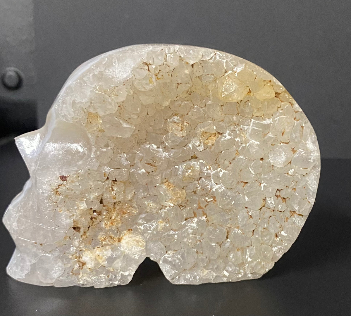 A carved skull-shaped flower agate and quartz crystal geode with rough, translucent pale quartz and natural golden-brown inclusions, photographed against a dark gray background.