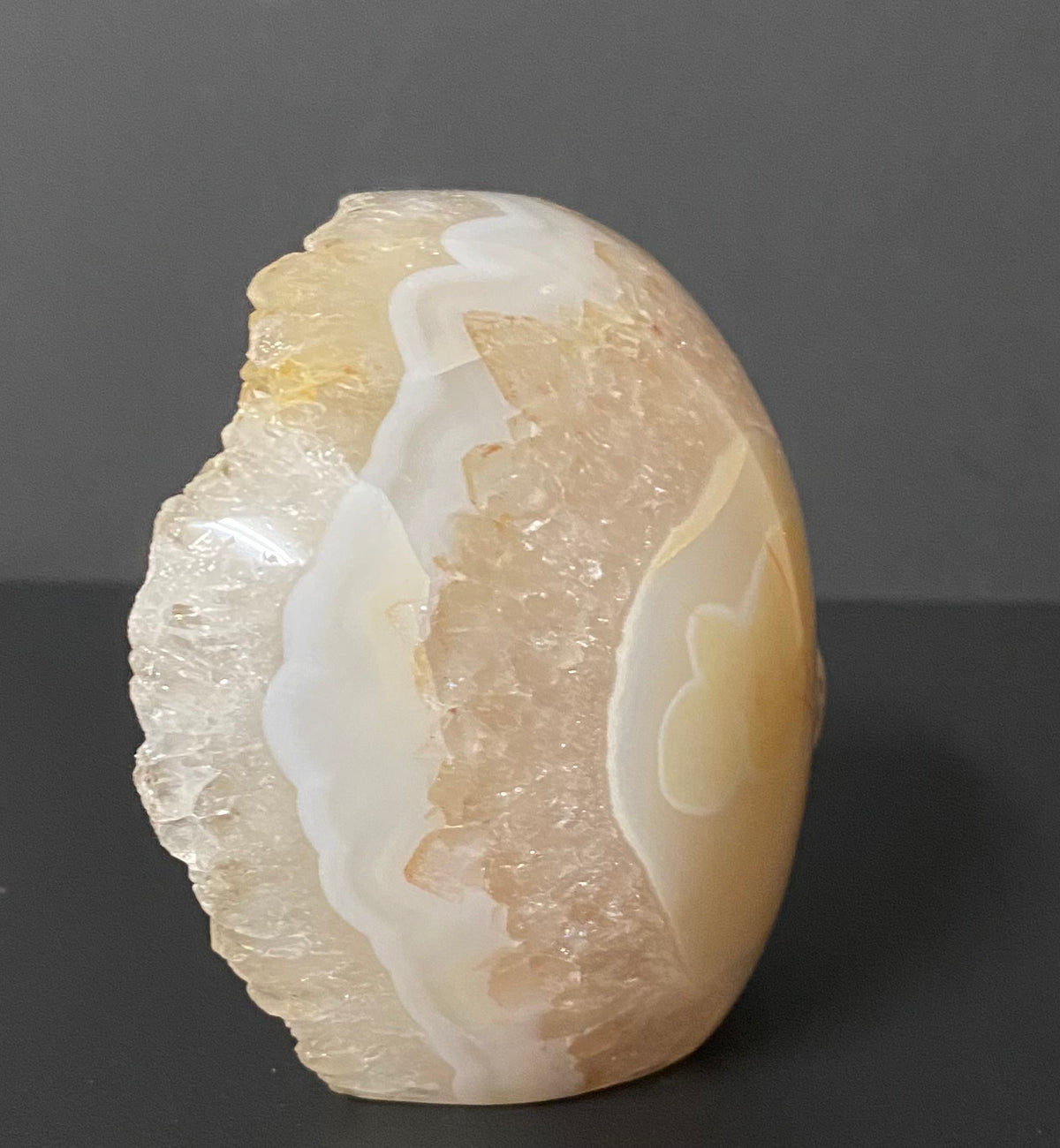 A polished oval flower agate and quartz crystal stone sculpture with natural banding in cream, white, and pale peach-beige, standing on a dark gray surface against a dark gray background.