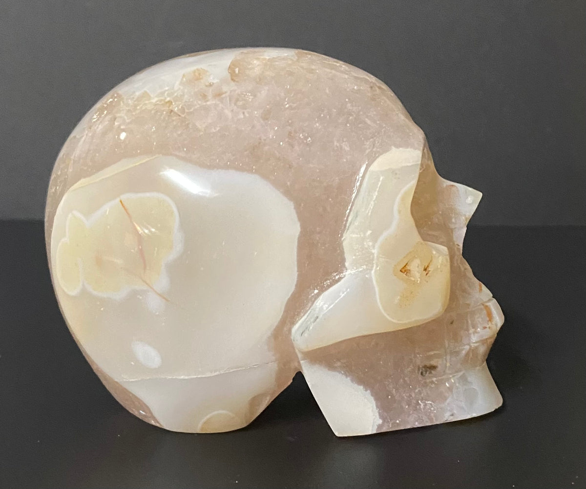 A carved natural flower agate and quartz crystal skull sculpture, showcasing milky white and soft peach-pink stone with natural inclusions, set against a dark gray background.
