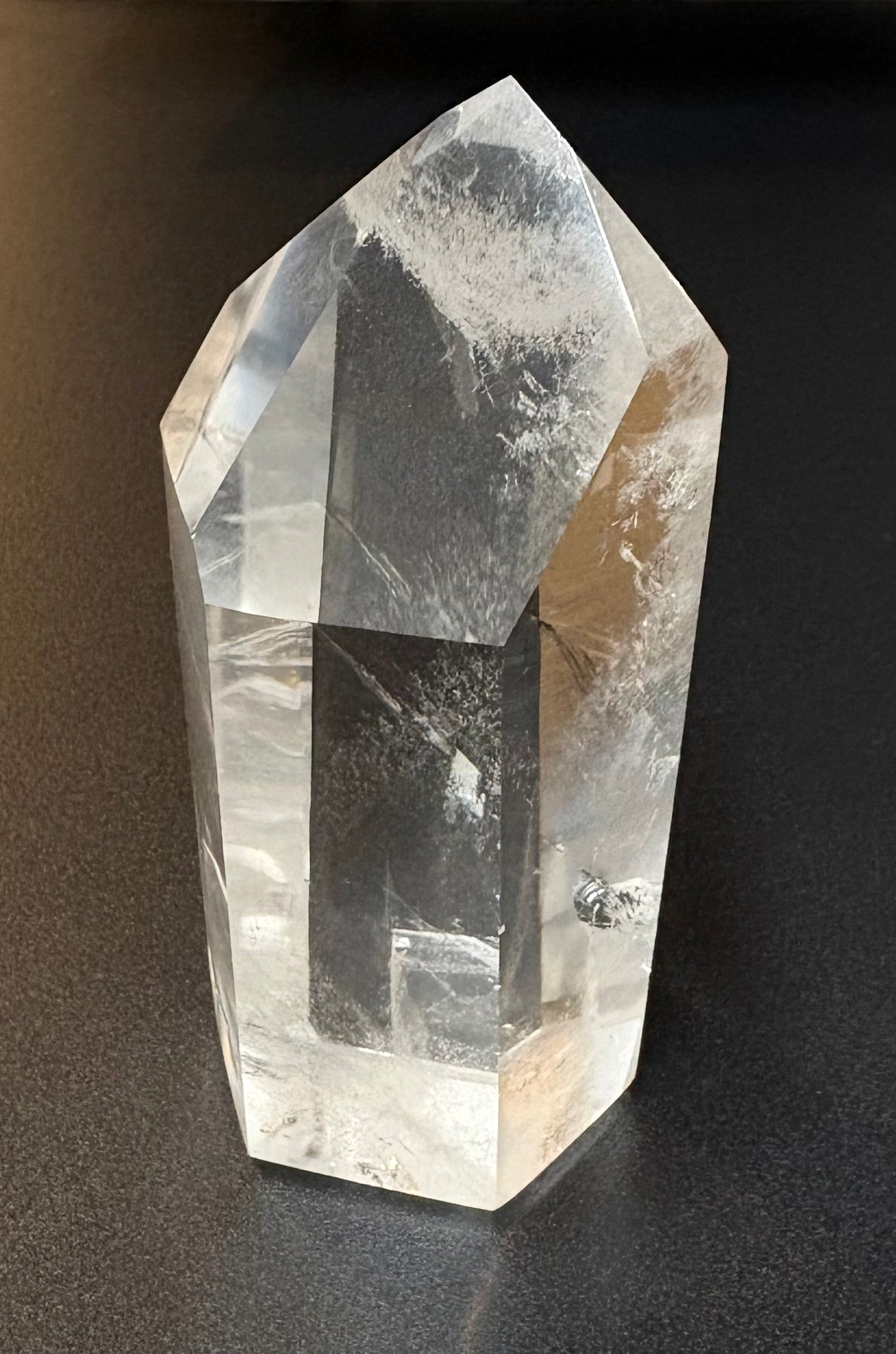 A clear quartz crystal point tower with natural inclusions and cloudy white patterns, standing on a dark gray surface.