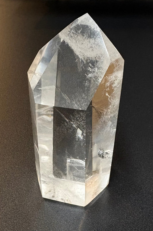 A clear quartz crystal point tower with natural inclusions and cloudy white patterns, standing on a dark gray surface.