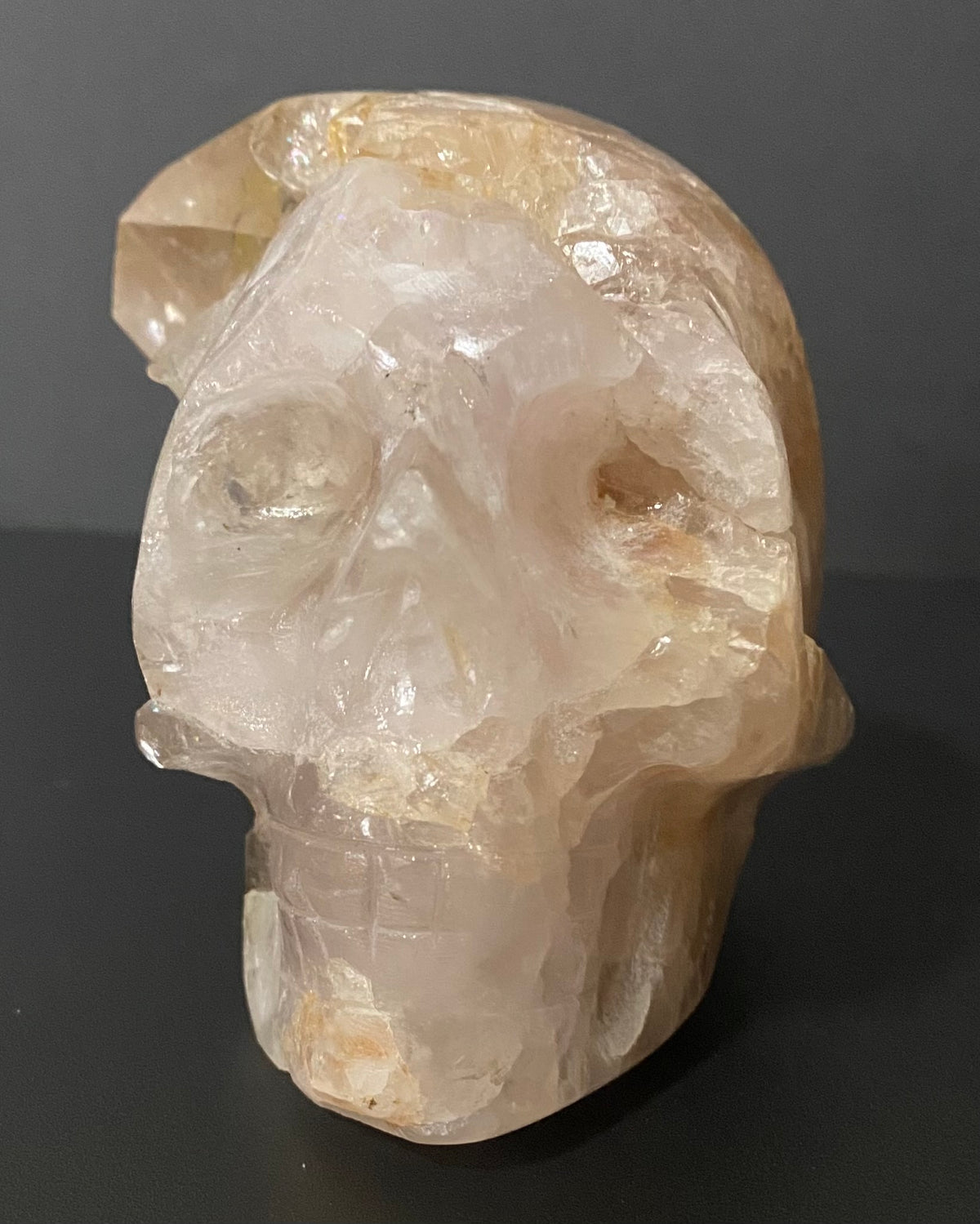 A carved crystal skull sculpture made from pale, translucent quartz with warm tan and cream tones, photographed against a dark gray background.