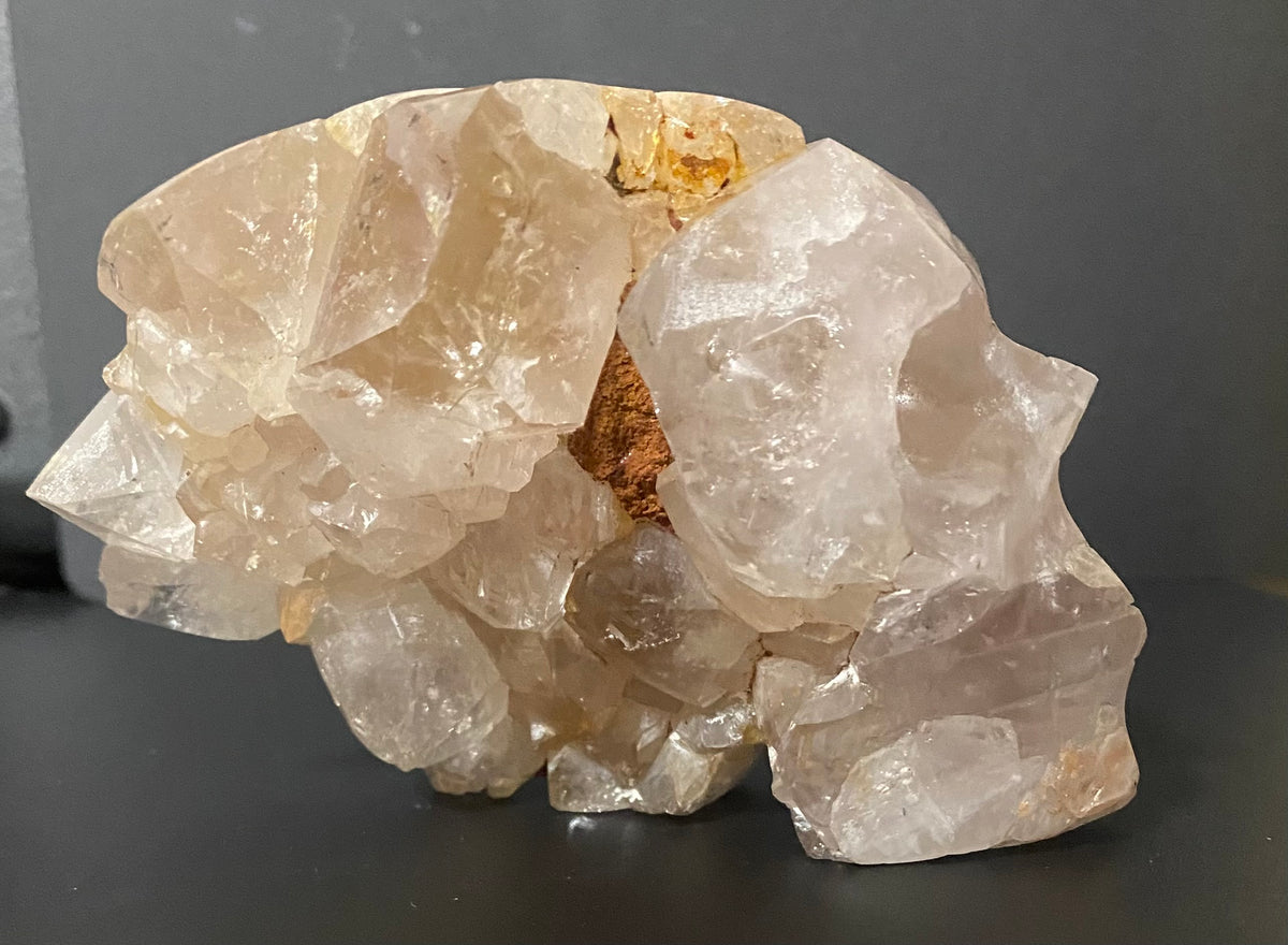 A raw crystal cluster specimen, primarily composed of translucent pale super seven quartz with a small exposed brown matrix, displayed on a dark surface against a dark gray background.