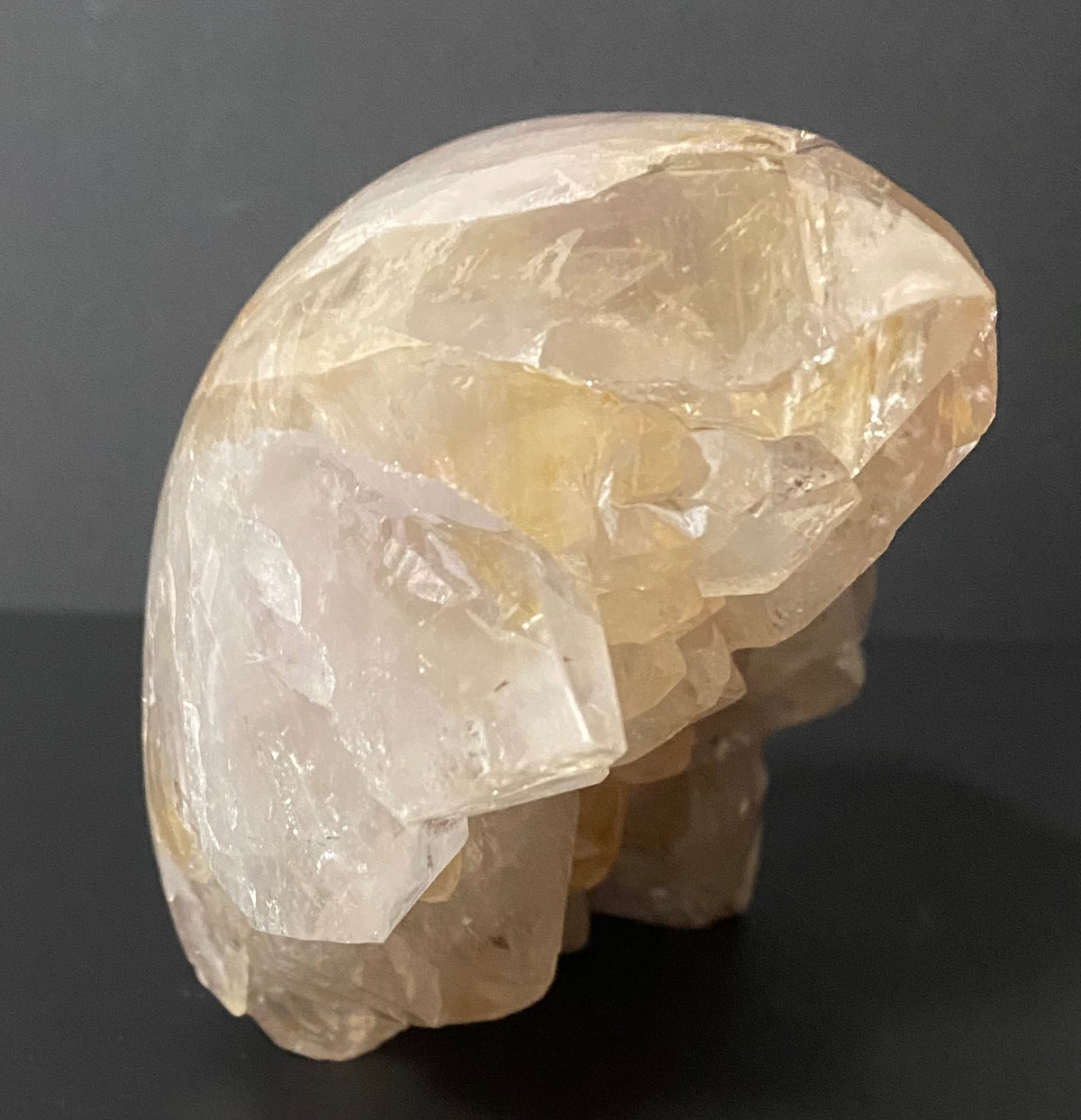 A raw, polished crystal skull specimen made of translucent super seven quartz, standing on a dark black surface against a dark gray background.