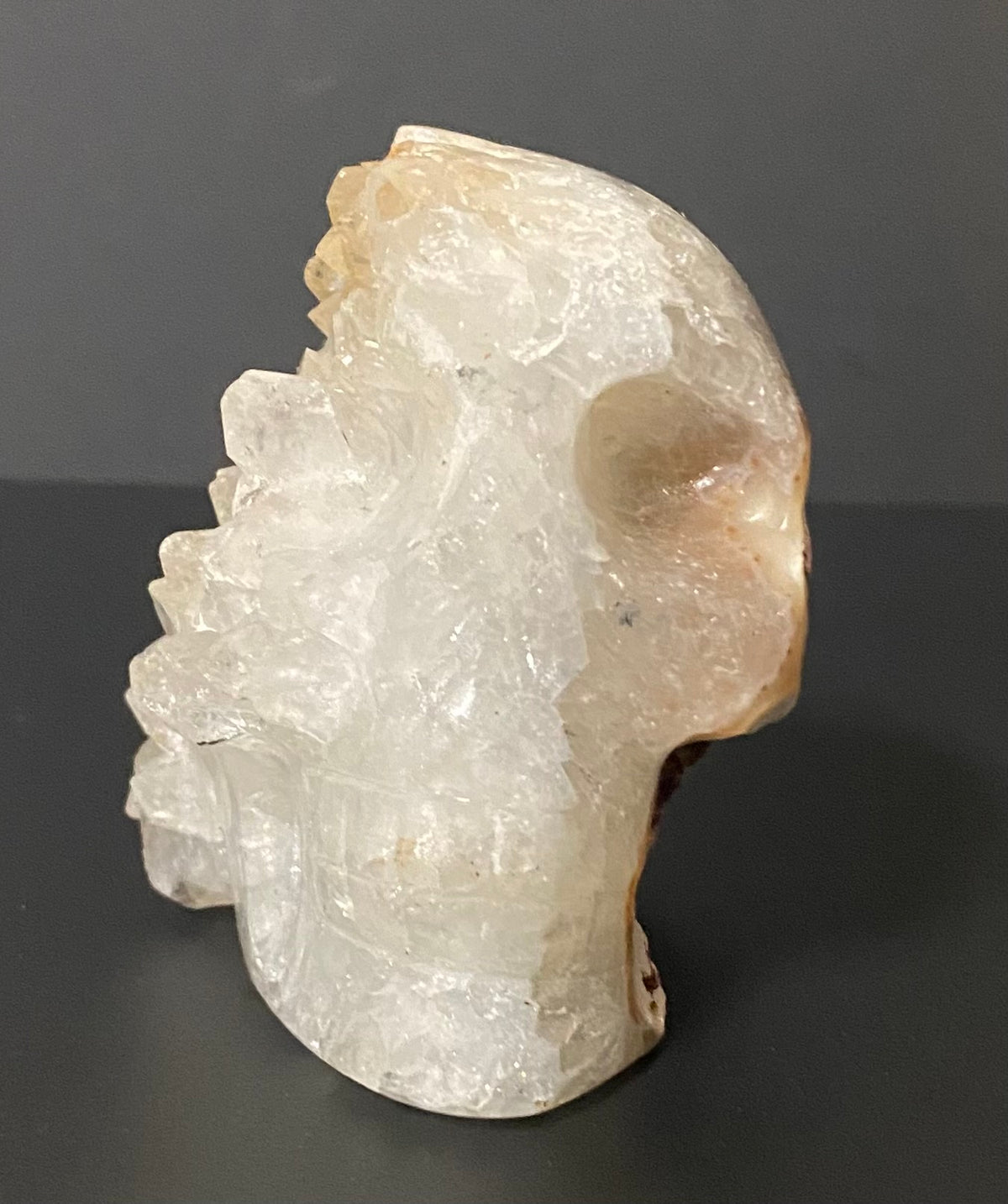 A carved crystal skull sculpture made from translucent white and pale amber Botswana agate and quartz stone, photographed against a dark gray background.