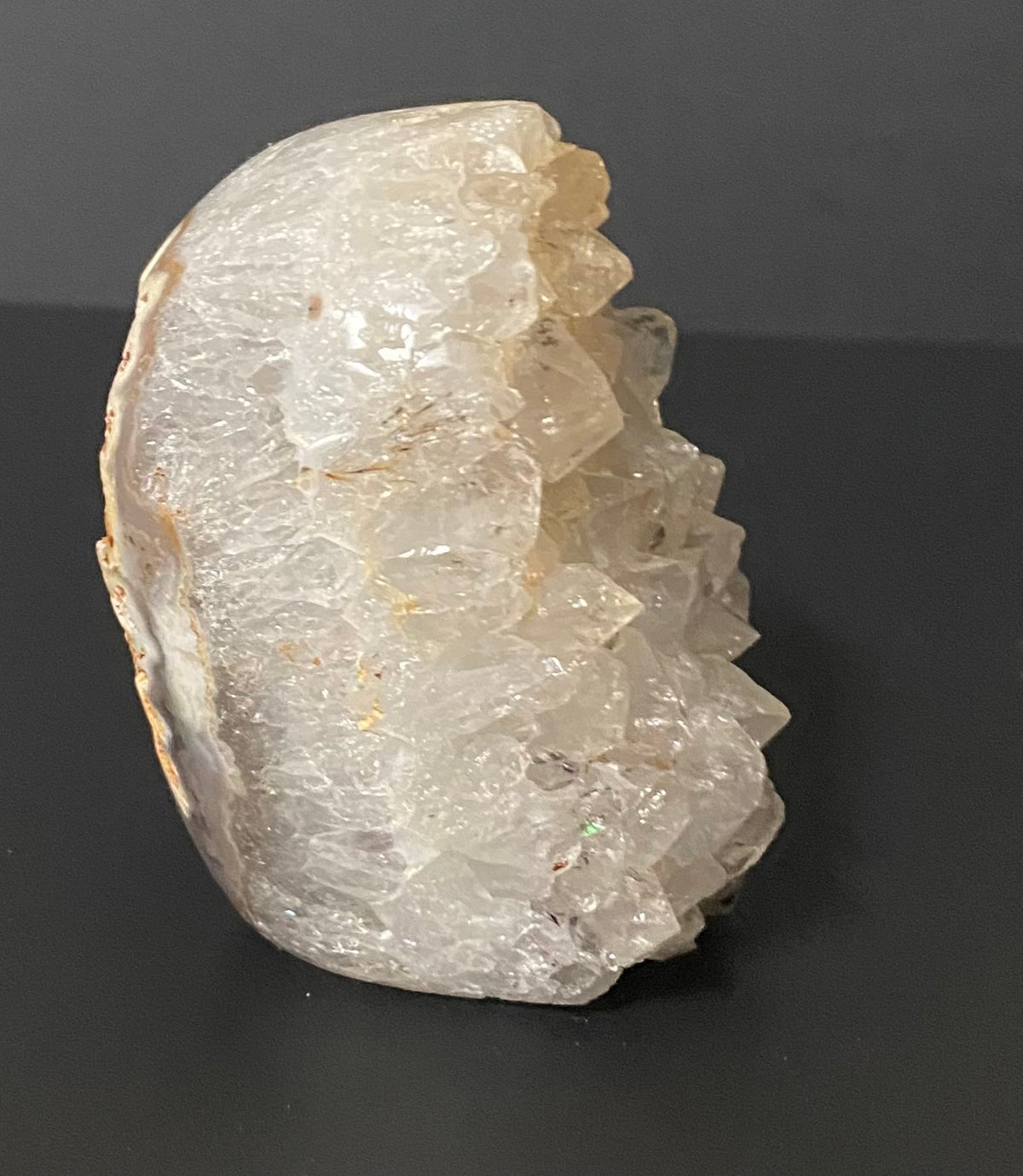 A raw crystal geode specimen with jagged translucent white and pale yellow quartz points and Botswana agate, standing upright on a dark gray surface against a dark gray background.