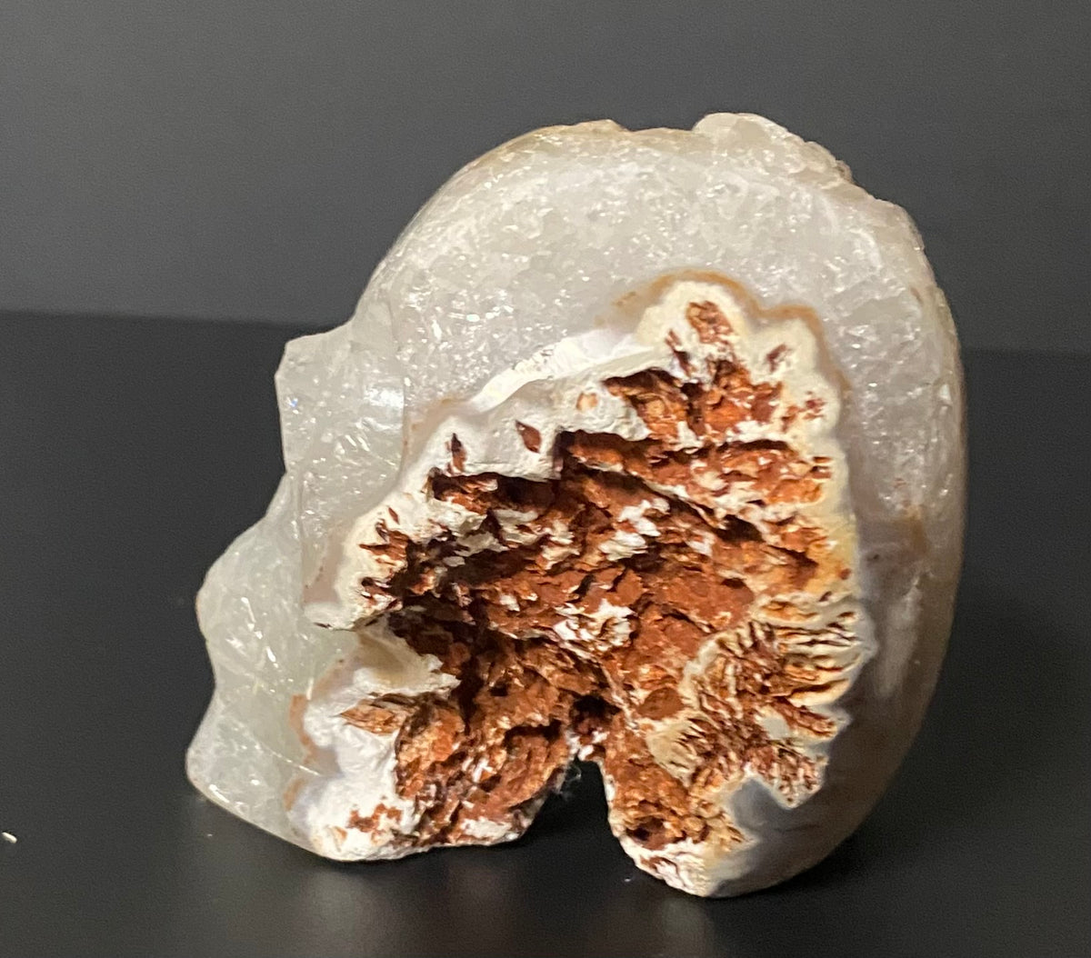 A polished natural mineral specimen with a smooth off-white outer surface and a textured, rust-brown crystalline interior, photographed on a dark gray surface and background. Botswana agate and quartz.