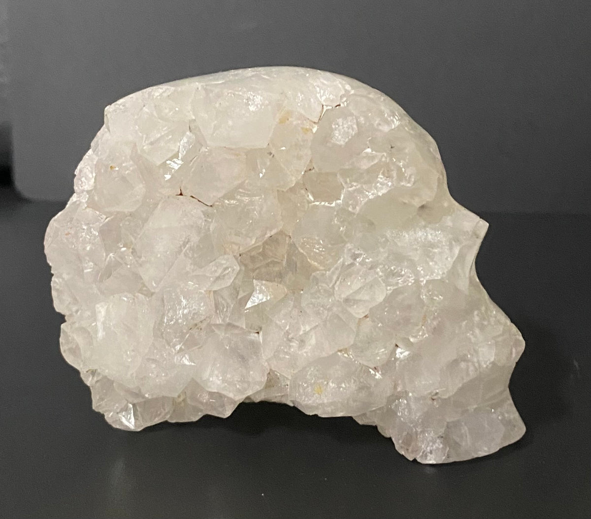 A raw, rough crystal cluster shaped like a stylized skull, made of translucent white quartz facets and Botswana agate, displayed on a dark gray surface.