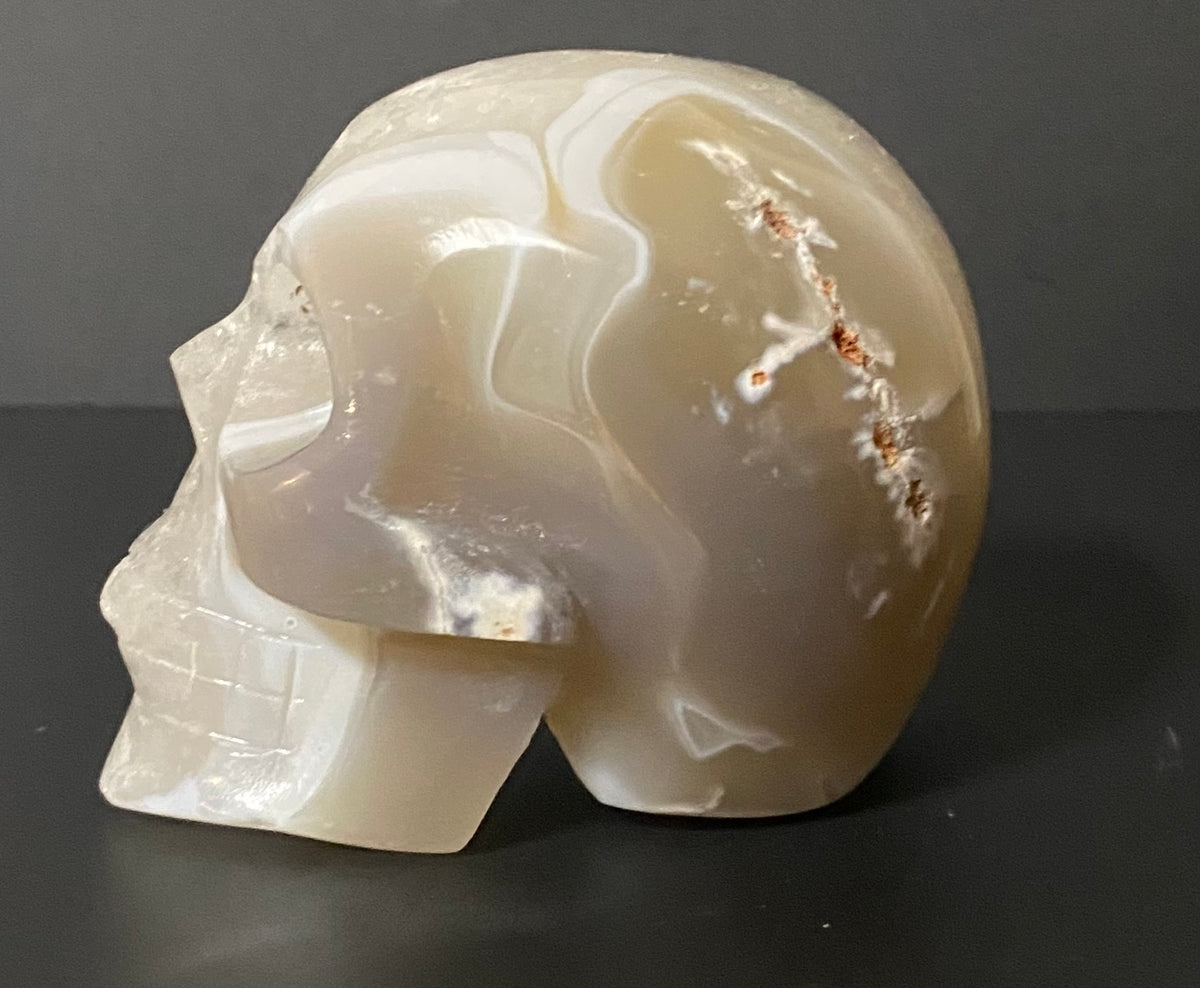 A carved Botswana agate and quartz skull sculpture with natural banding and reddish-brown inclusions, photographed on a dark gray background.