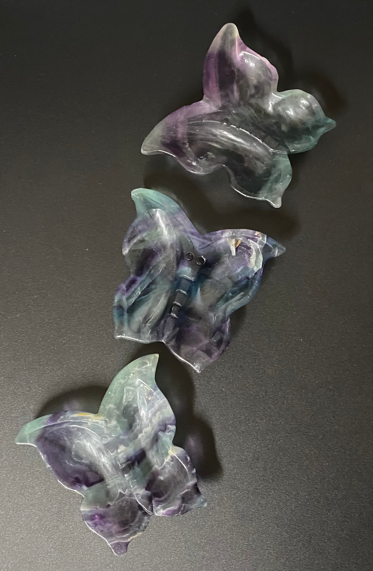 Three translucent fluorite butterfly-shaped decorative pieces with marbled purple, blue, and green tones arranged diagonally on a dark gray background.