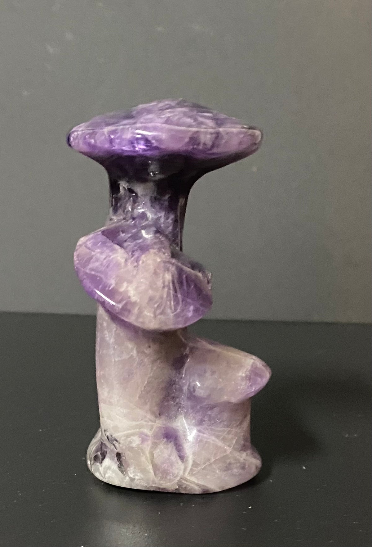 A small polished purple amethyst sculpture shaped like a stylized mushroom with three mushroom caps, standing on a dark surface against a gray background.