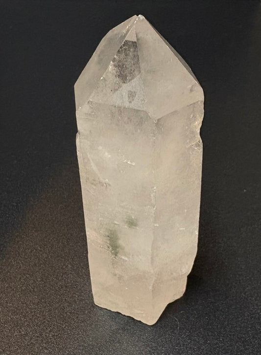 A natural clear quartz crystal point with sharp geometric facets, standing upright on a dark textured surface.