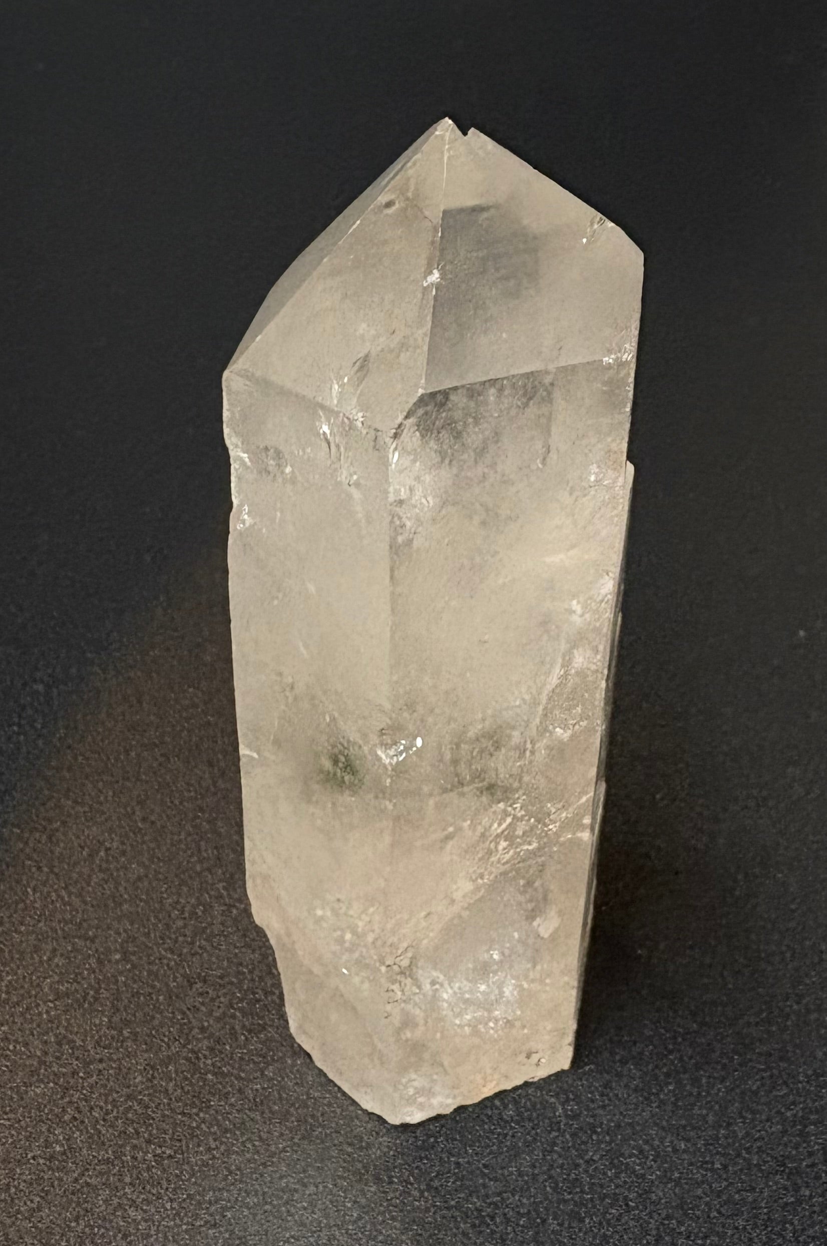 A natural clear quartz crystal point specimen standing upright on a dark gray surface.