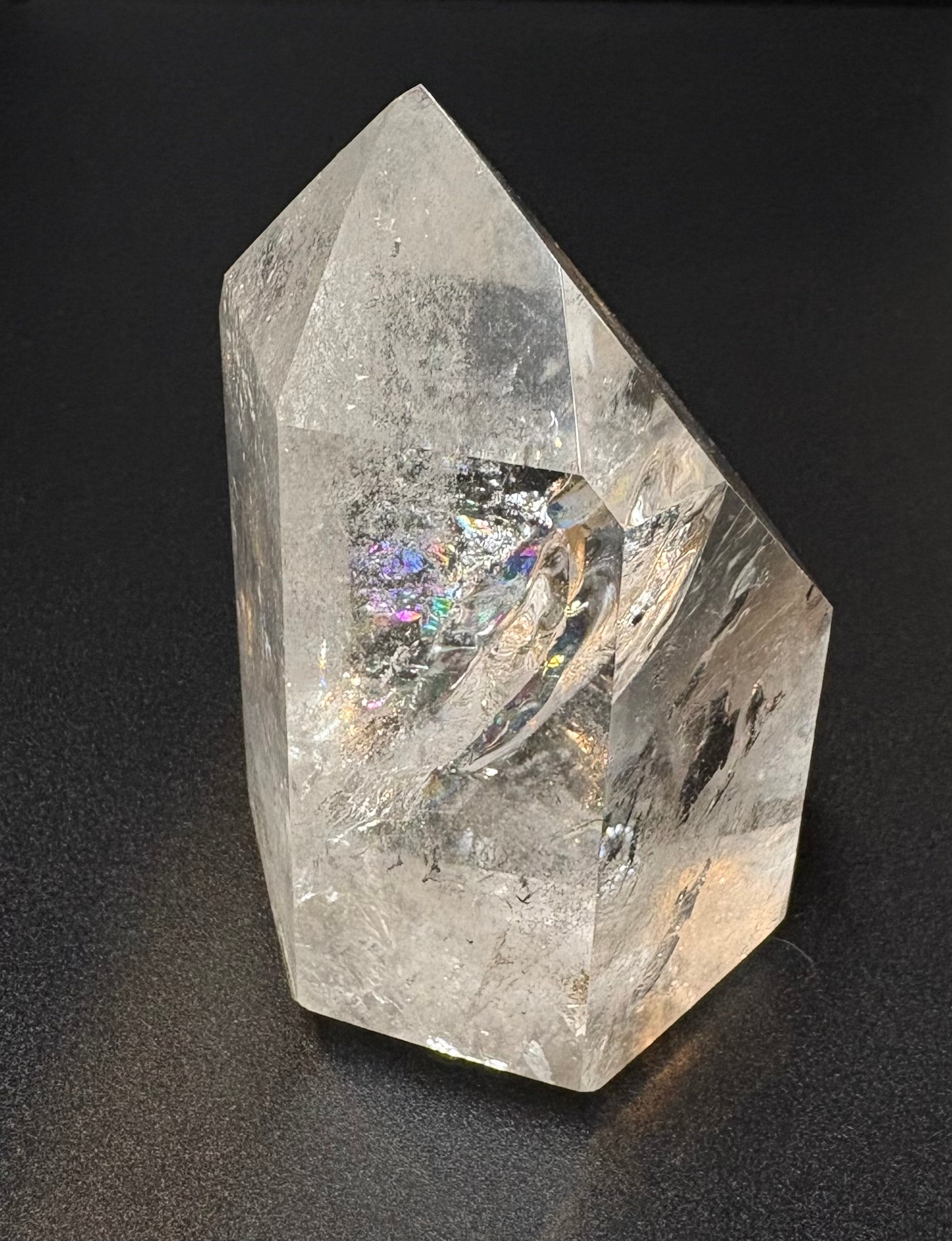 A natural clear quartz crystal point with internal inclusions and a subtle iridescent rainbow reflection, set against a dark textured background.
