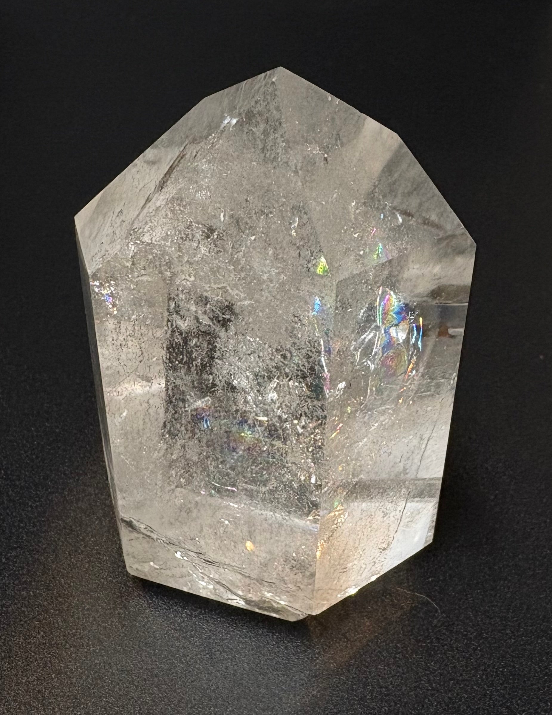 A clear faceted crystal point, likely quartz, with natural internal inclusions and subtle iridescent highlights, photographed on a dark black background.