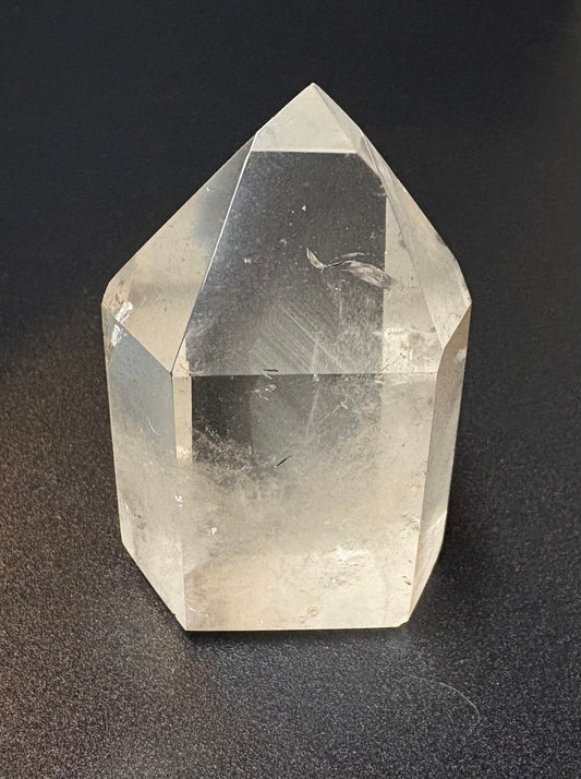 A clear pointed crystal tower or geode specimen with faceted sides, photographed upright on a dark textured surface.
