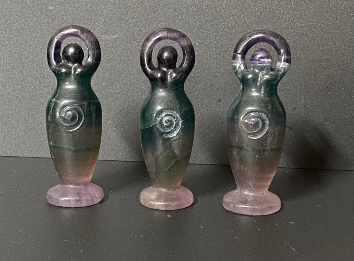 Three small fluorite goddesses shaped like stylized female figures with raised arms and spiral details on their torsos, standing on round bases against a dark gray background.