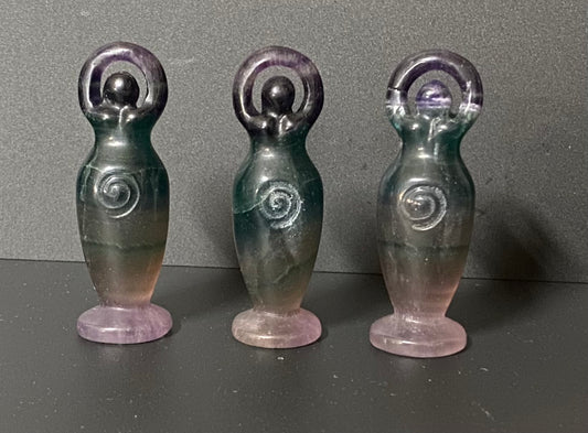 Three small fluorite goddesses shaped like stylized female figures with raised arms and spiral details on their torsos, standing on round bases against a dark gray background.